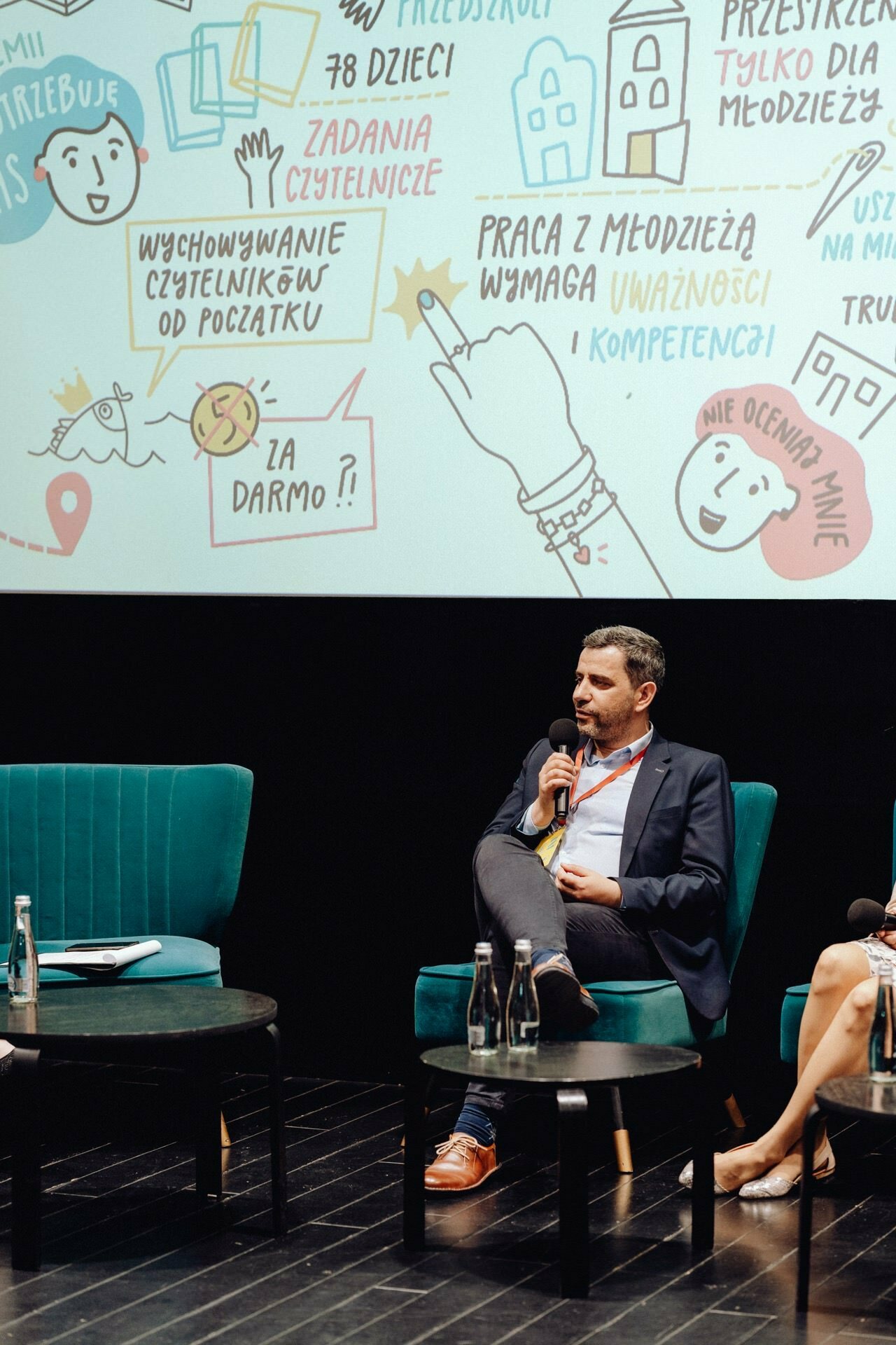 A man in a suit sits on stage and speaks into a microphone. In the background is a projection screen with colorful illustrations and Polish text. In front of him are two turquoise armchairs and a round black table with water bottles, perfectly captured by the photographer for the event.  