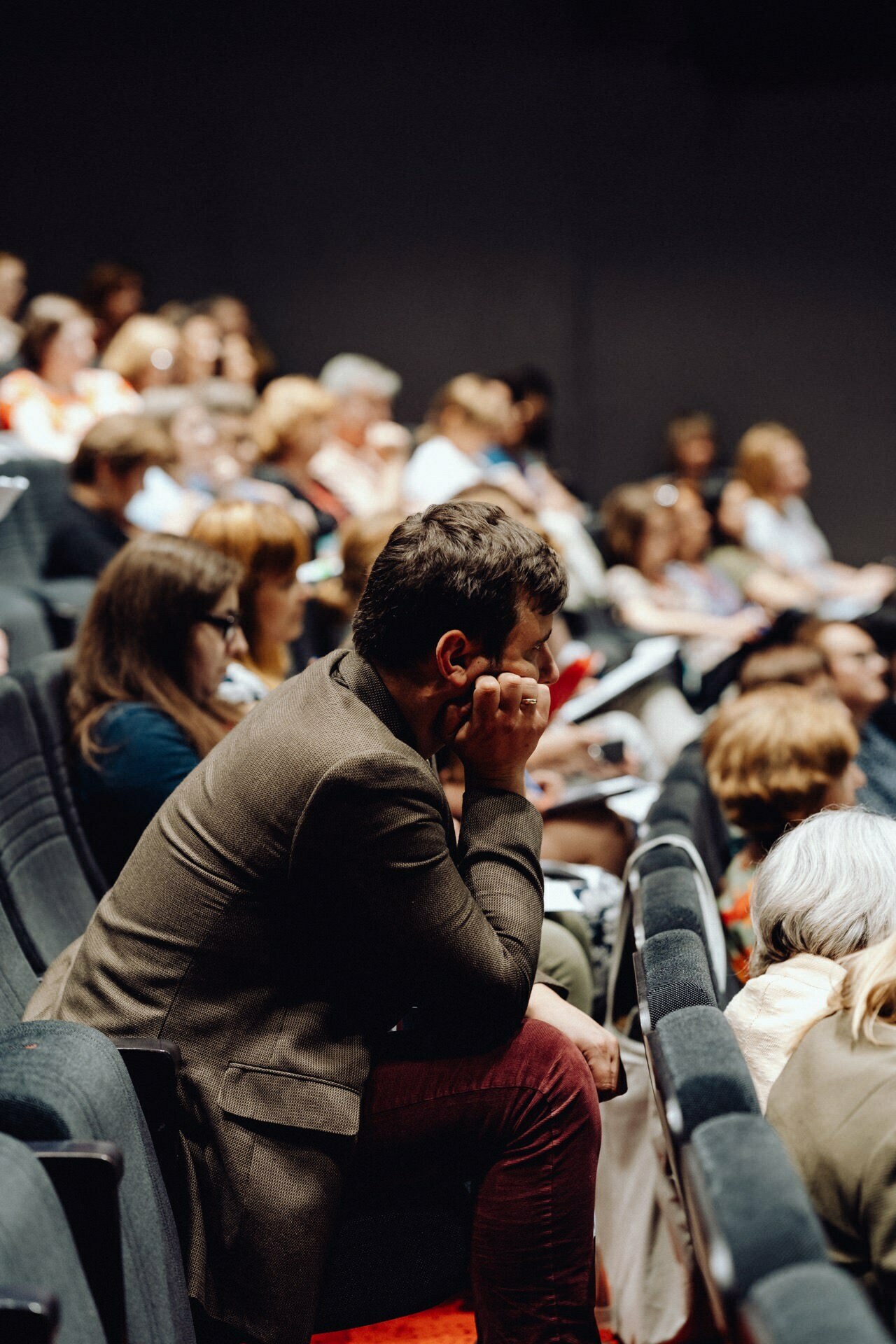 A group of people sitting in a dimly lit auditorium listening intently to a presentation. One person in the foreground, wearing glasses and a brown jacket, rests his chin on his hand in a reflective pose. This moment was captured by a photographer from Warsaw, showing more people in the background.  