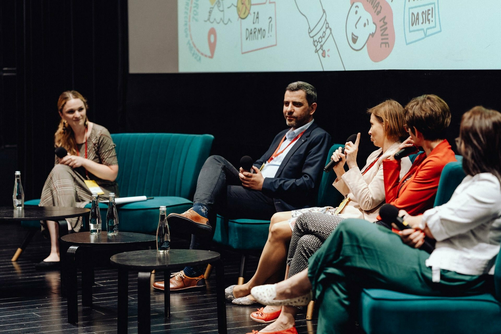 On a stage with microphones sit five people engaged in a panel discussion. Behind them is visible a projector screen displaying the illustrated content. Three green armchairs have been set up on the stage, and tables with bidons have been set up in front of the participants - the perfect stage for a photo-op of the event.  