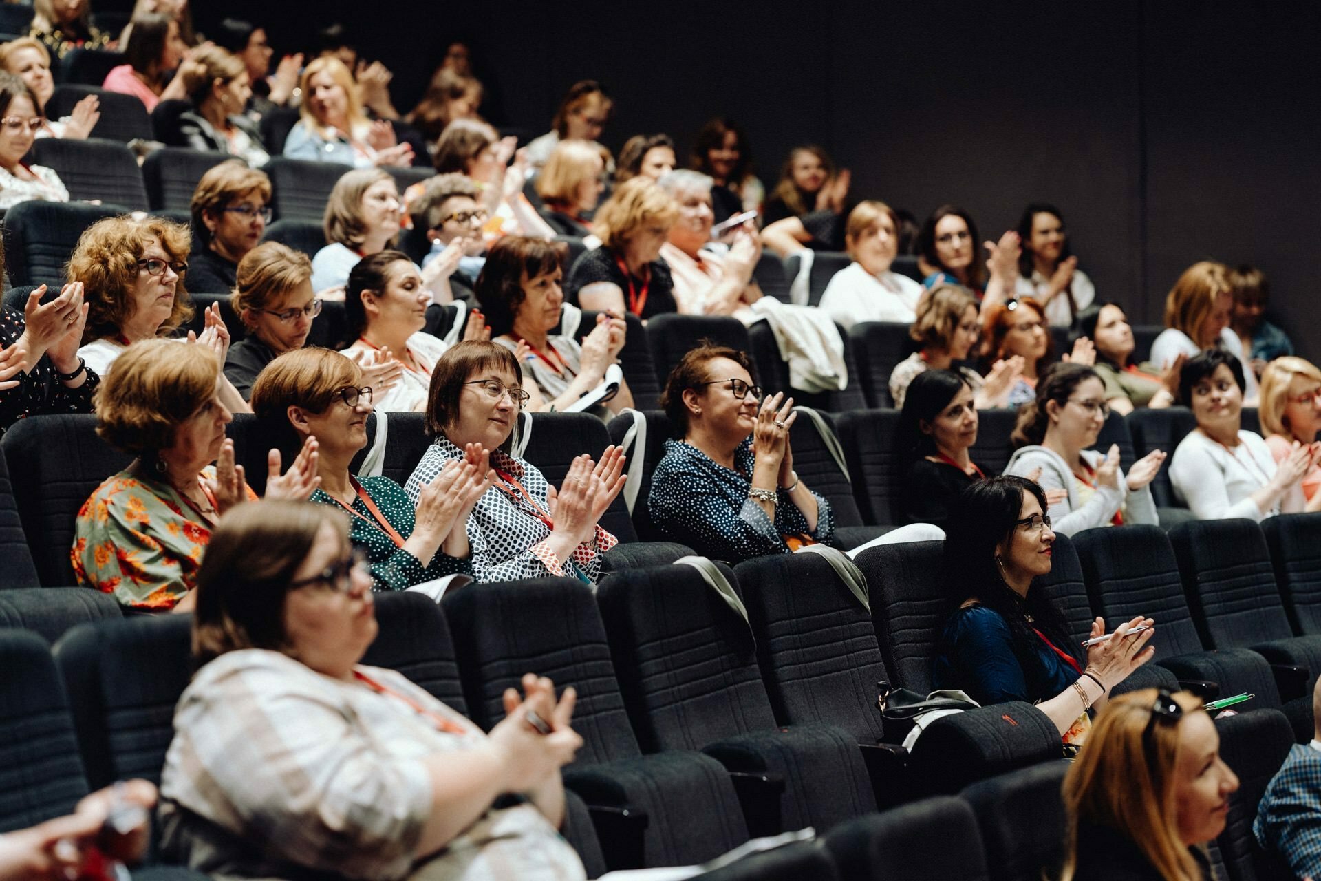 A large group of people, mostly women, sitting in a theater-like room. They clap and seem engaged, facing forward. Some are smiling, wearing badges and taking notes. The atmosphere seems lively and attentive - perfect for a photographer at an event in Warsaw, Poland.   