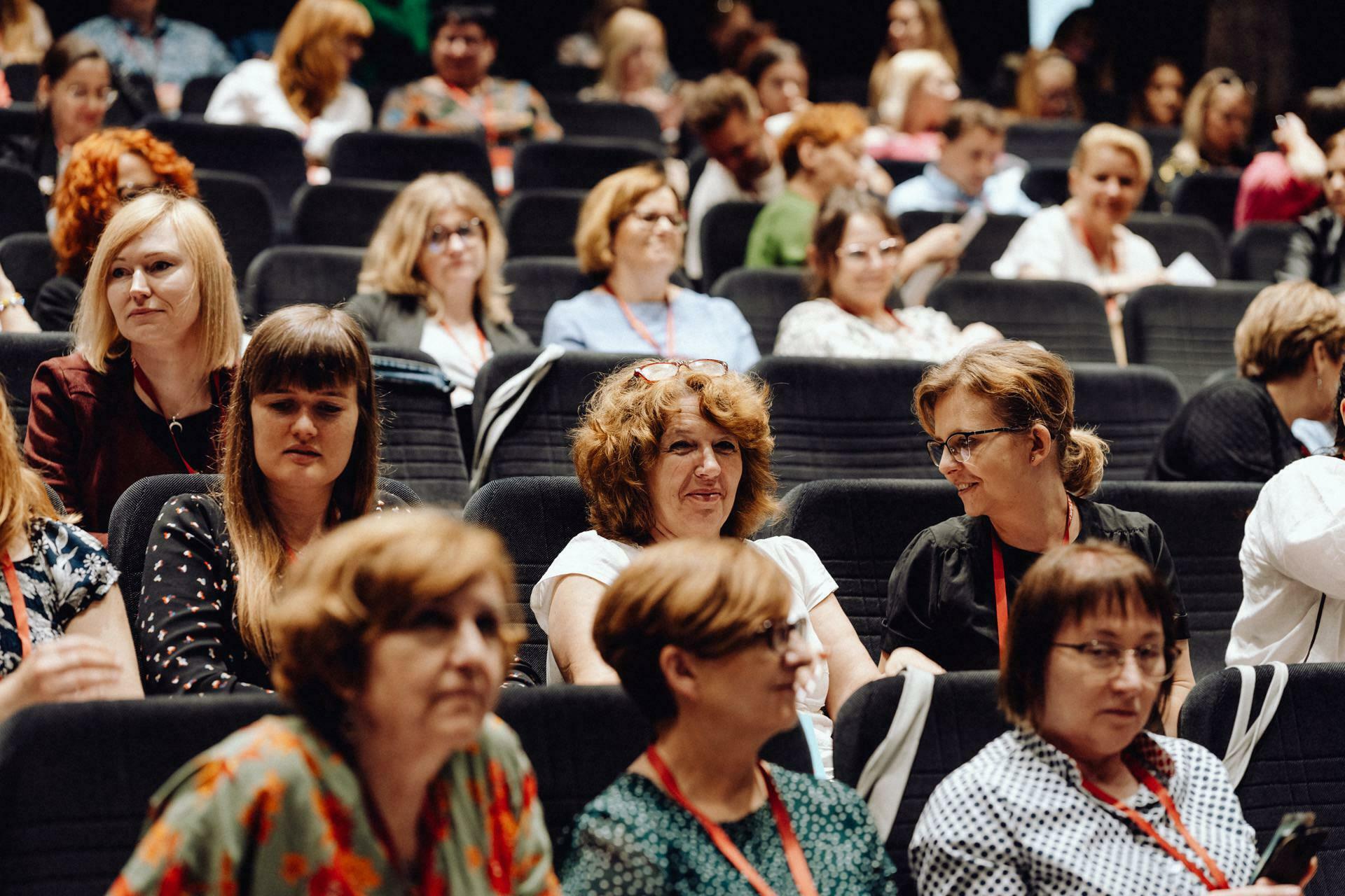 A large group of people, mostly women, sit in rows of auditorium chairs. Many are busy talking, others are looking ahead. Most wear casual clothes and have name badges on lanyards around their necks. This captivating photo report of the event perfectly captures the vibrant atmosphere.   