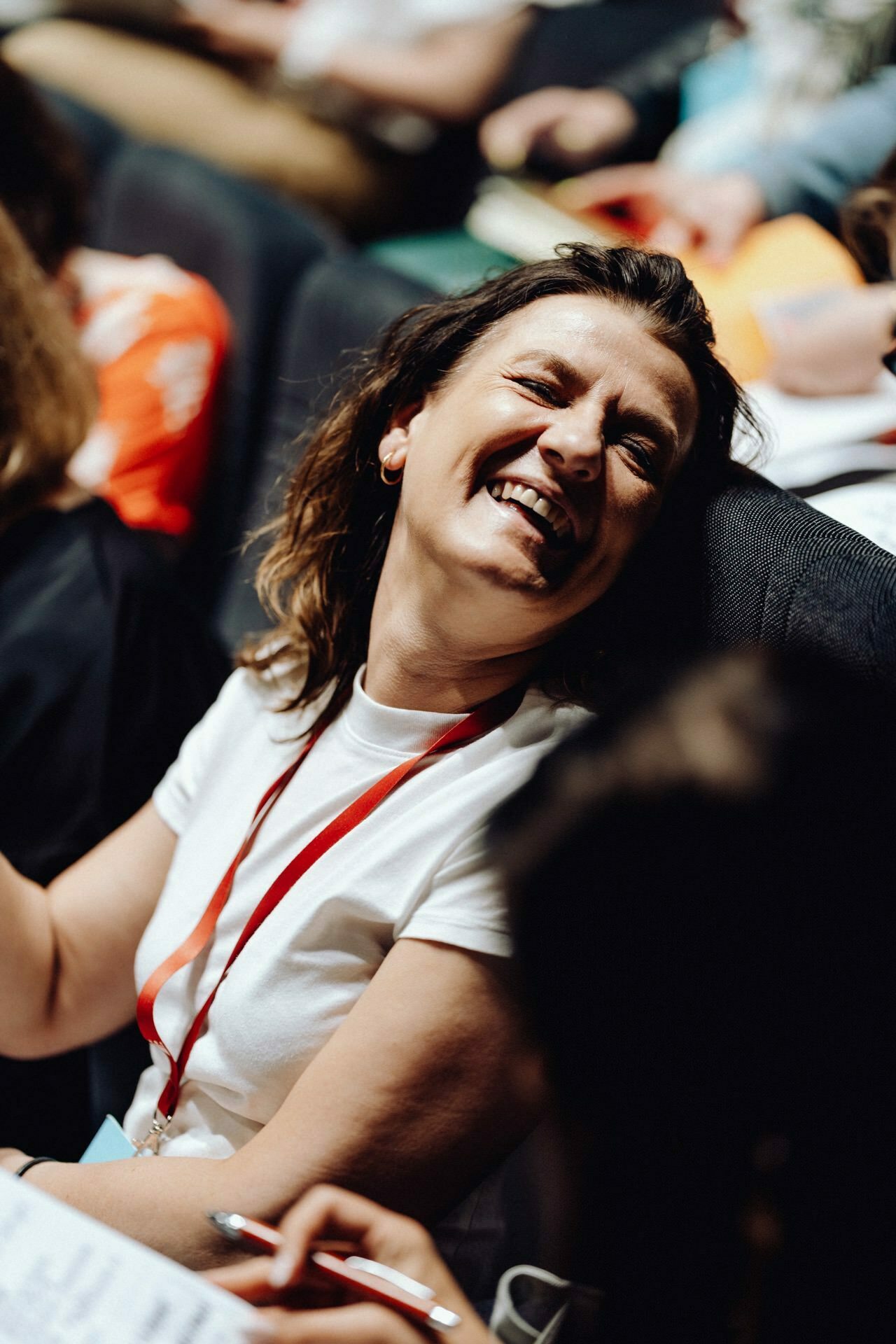 A person with shoulder-length hair, wearing a white shirt and red lanyard, laughs while sitting among the crowd, showing joy and engagement. In the background, blurred people and elements can be seen, suggesting an event or environment captured by the photographer at the event. 