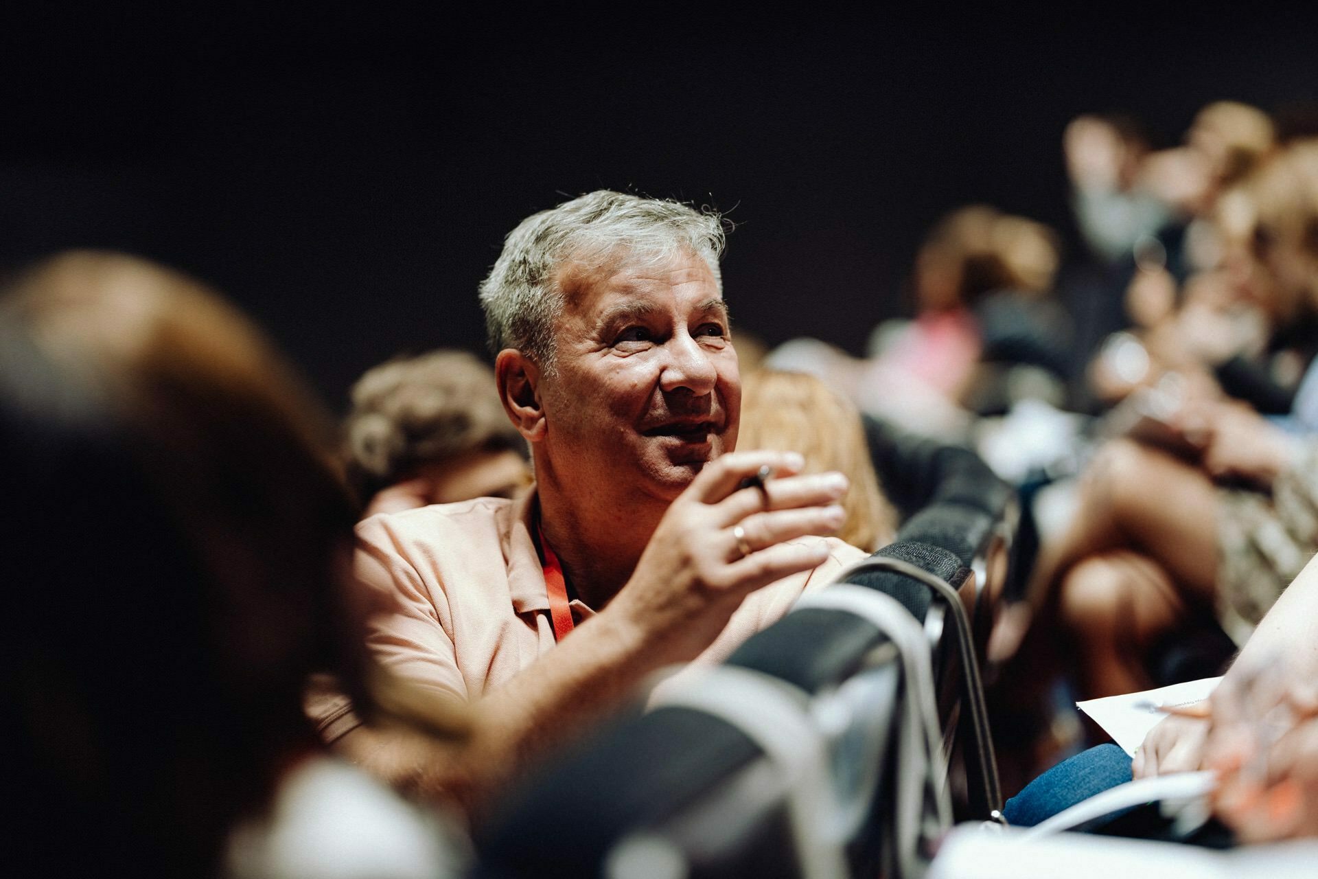 A man with gray hair, wearing a coral-colored jacket, is sitting in an auditorium among a crowd of people. He is smiling and looks as if he is busy talking to someone next to him. The background is blurred, showing other attendees sitting nearby - perfect for a photo essay of the event.  