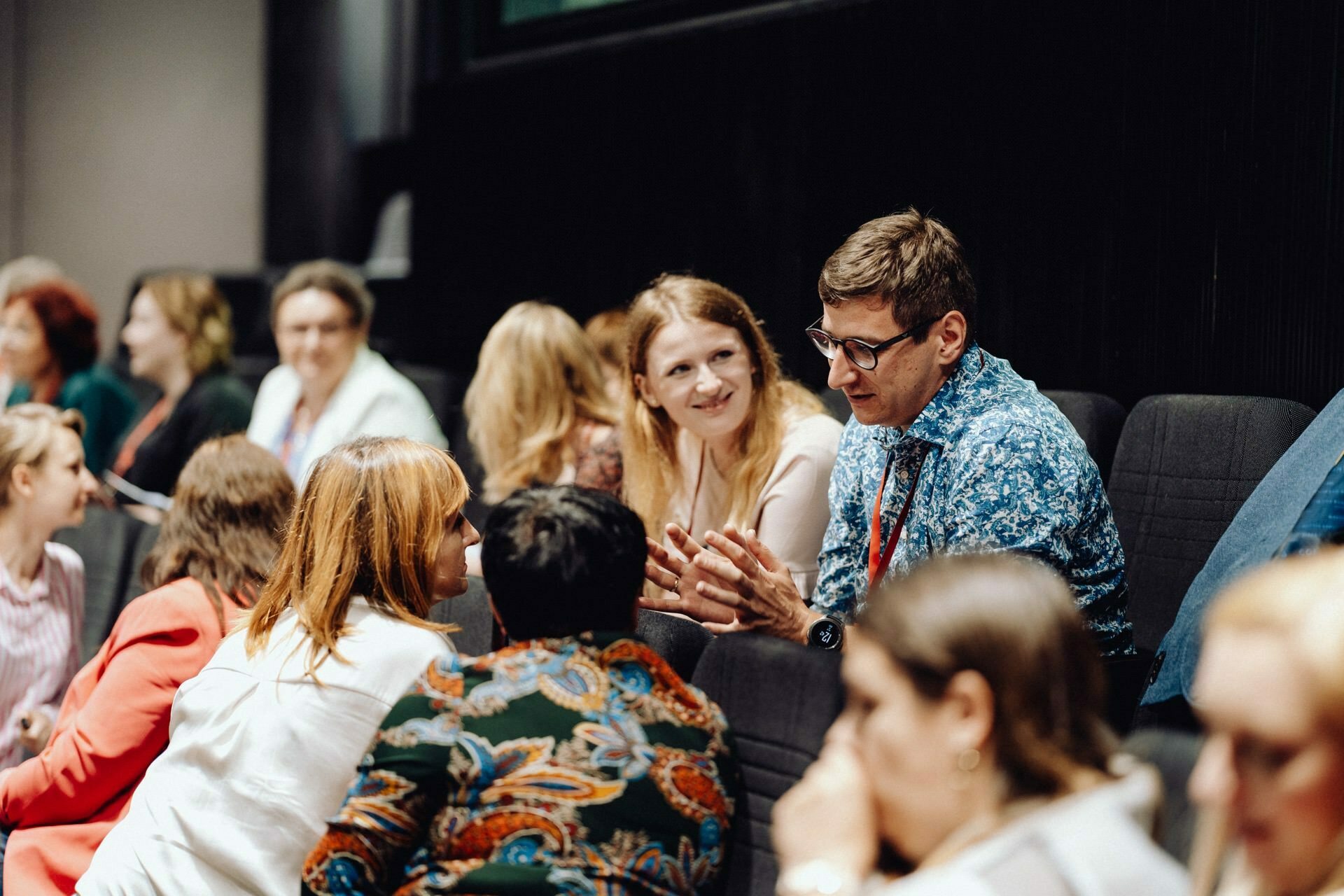 In an auditorium-like seating arrangement, people hold animated conversations. A man in a patterned blue shirt and glasses warmly chats with a red-haired woman in a white blouse. The vibrant scene of interaction was beautifully captured by event photographer Warsaw, documenting the memories for the event photo report.  