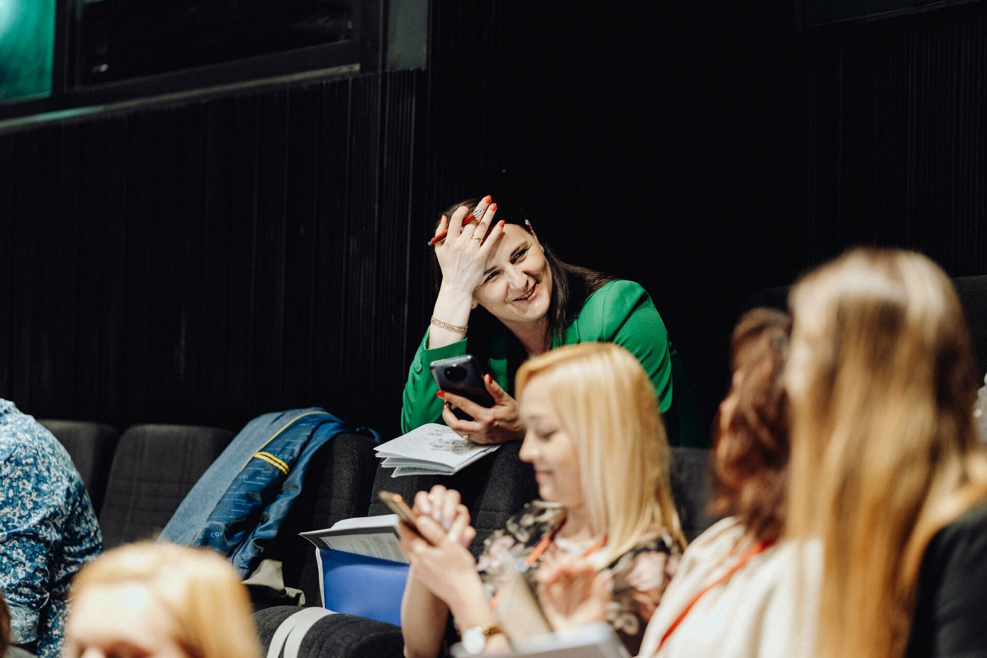 A woman in a green blouse, holding a phone and notepad, smiles and rests her head on her hand, looking at another person. Captured during an event photo shoot by a photographer for the event, she sits among a group of people in what resembles a lecture hall or auditorium. 