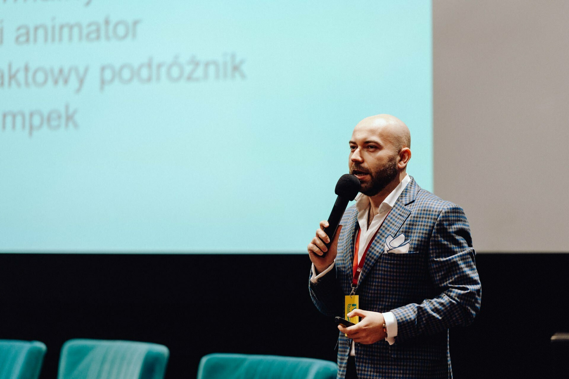 On stage, a man in a plaid jacket speaks into a microphone. Behind him is a presentation screen with text in a foreign language. Blue chairs are set up next to him, capturing a photo moment of the event.  