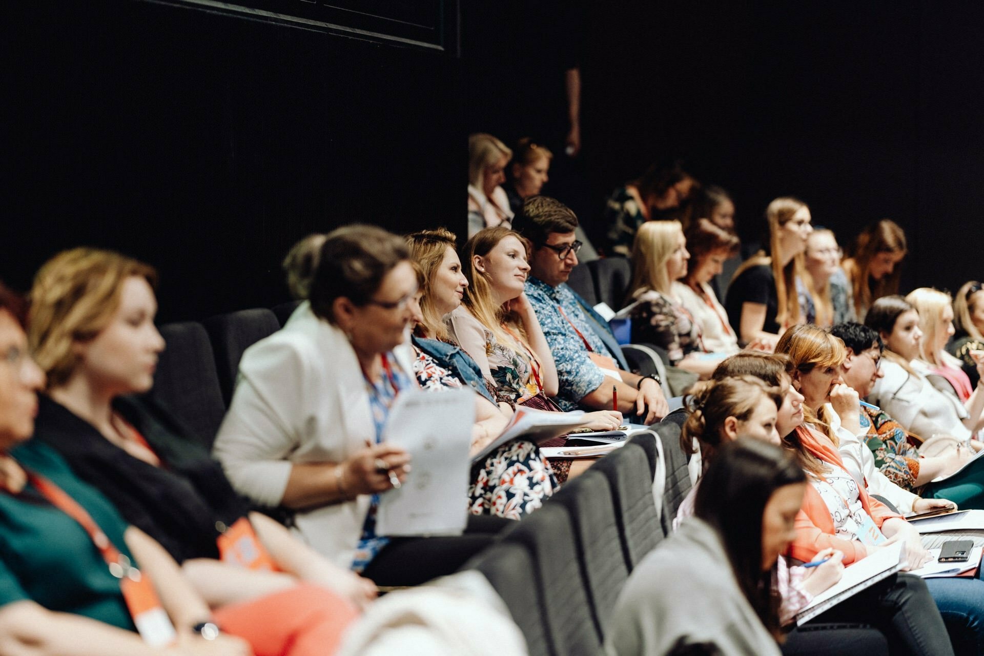 A diverse group of people sit attentively in a darkened room, focused on the presentation. Some are taking notes and holding papers, while others are listening intently. The mood suggests a professional or educational event, perfect for a photo essay of the event captured by a photographer from Warsaw.  