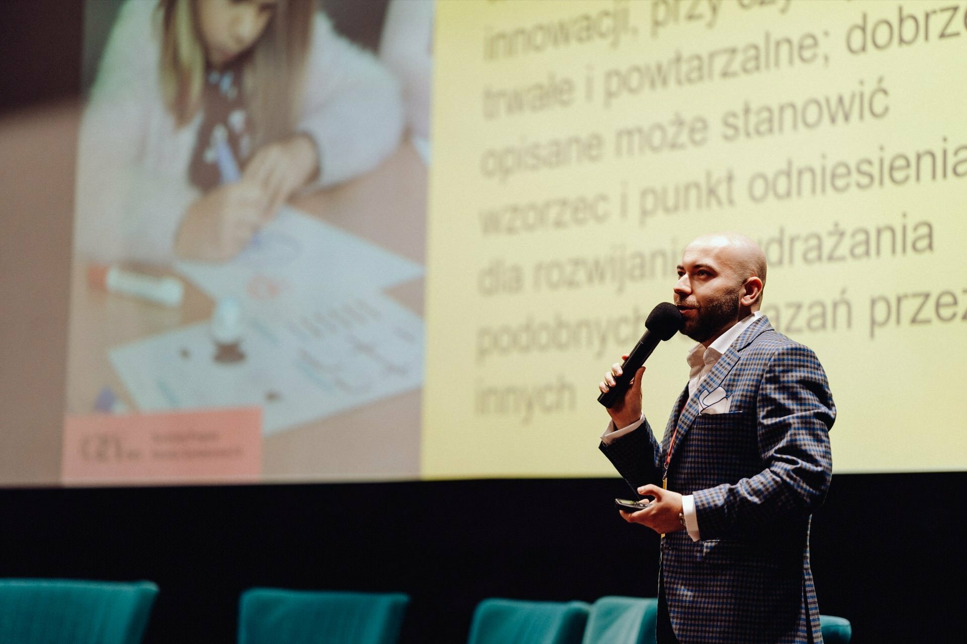 On stage, a man in a plaid suit speaks into a microphone. Behind him is a large screen showing a child drawing on paper with markers, accompanied by text in Polish. The scenery is reminiscent of a conference or presentation, captured as part of a photo report of the event by a photographer from Warsaw.  