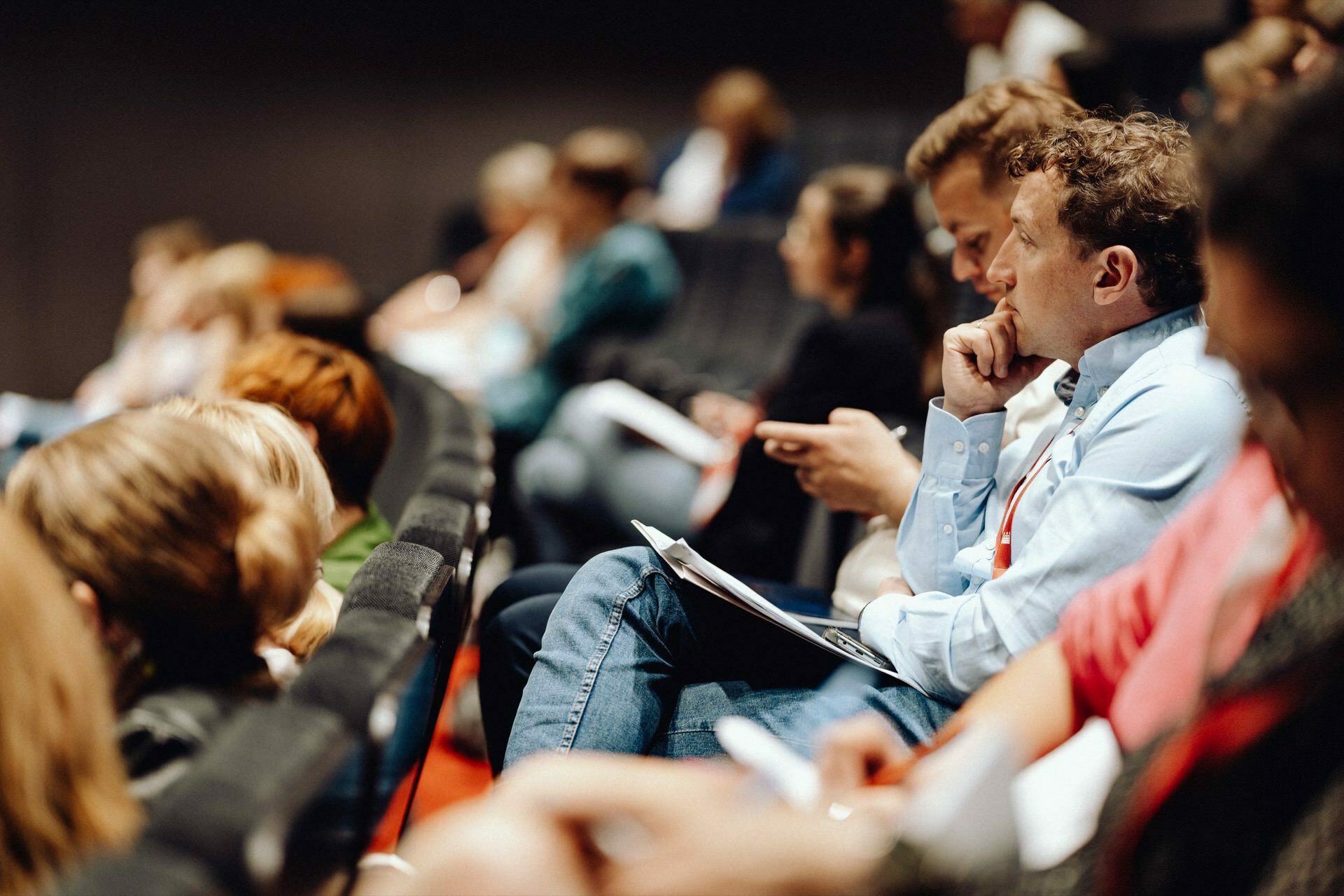 A diverse group of people sitting in rows in the auditorium. Some are taking notes, others are listening intently to the presentation. In the center of attention is a thoughtful man in the foreground with a pen to his chin, captured by a photographer from Warsaw providing photo coverage of the event.  