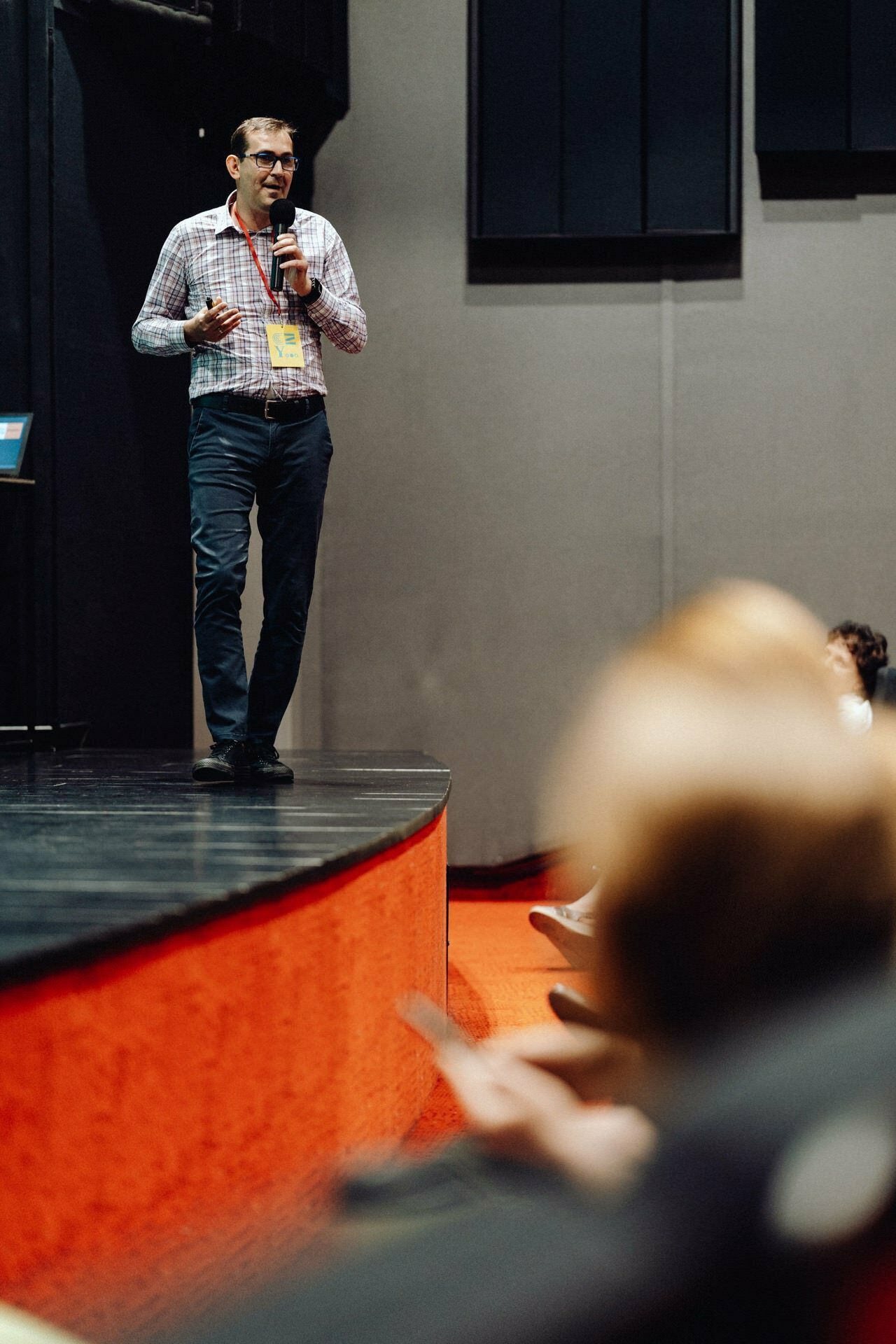 A person stands on stage dressed in a checkered shirt and black pants, holding a microphone in one hand and a device in the other, presumably giving a presentation. The background is a gray and red audience with part of the audience in the foreground, perfectly captured by the photographer for the event. 