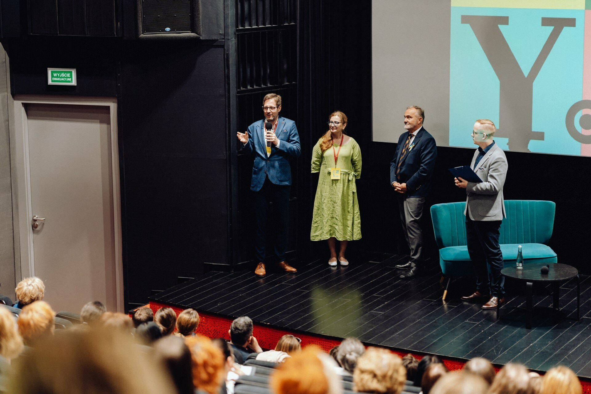 A group of four people stands on the stage in front of the audience. One person speaks while holding a microphone. On the stage is a blue chair and a small round table, and behind them is a screen displaying large letters. An event photographer warszawa captures the moment as the audience watches intently.   