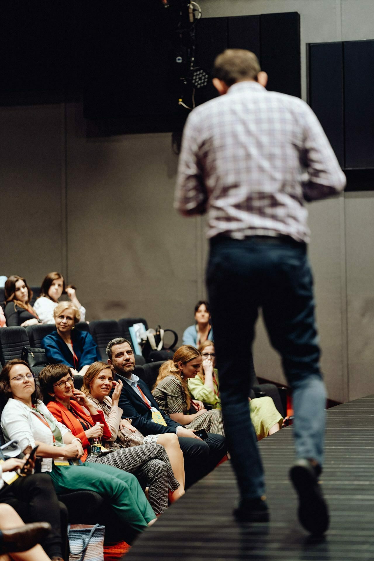 A speaker dressed in a checkered shirt and jeans appears on stage before an audience. Attendees, sitting in rows, watch the speaker intently. This in-house conference or seminar is expertly documented by a photographer for the event, who captures every moment for a photo report of the event.  