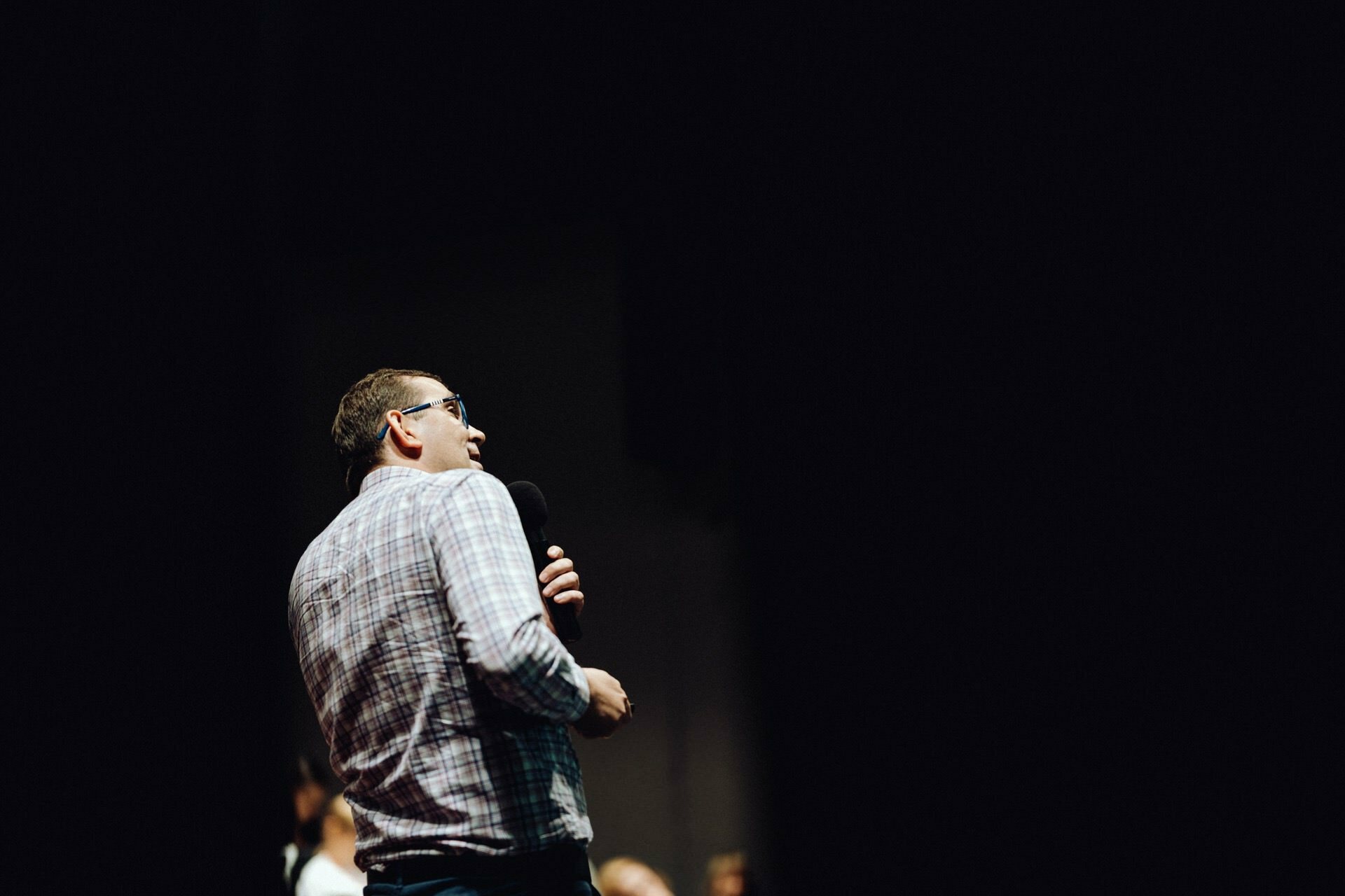 A man in a checkered shirt holds a microphone and speaks to an audience at an event. The background is dark, the man is illuminated, placed slightly off-center and facing right. Perfect for event photographer Warsaw to capture moments.  