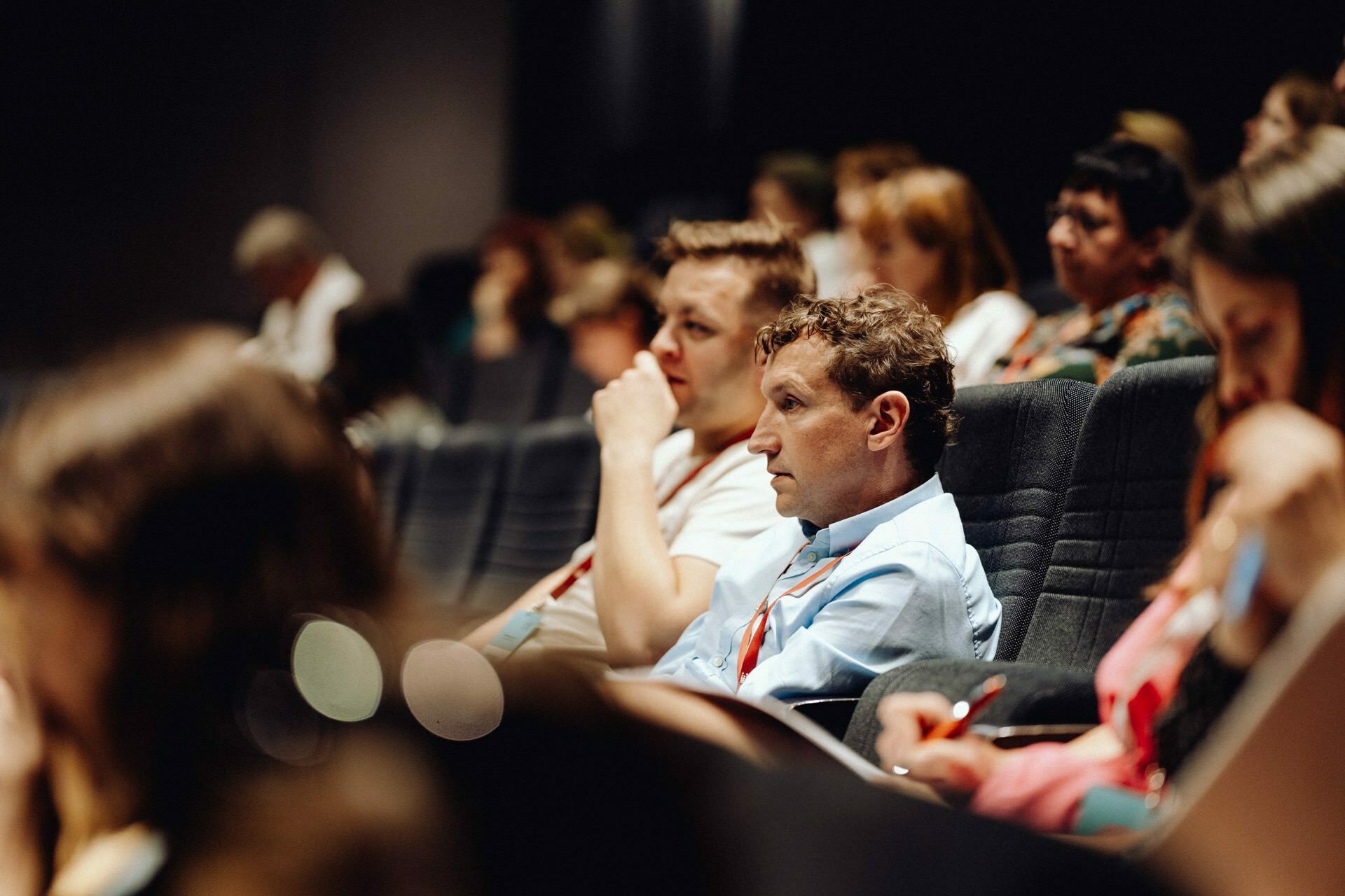 A group of people sit attentively in a dark auditorium, focused on an unseen speaker or presentation. They sit in rows, and some have notebooks and pens. The lighting is dim, emphasizing their focus - perfectly captured by the photographer at the event for a detailed photo report of the event.  