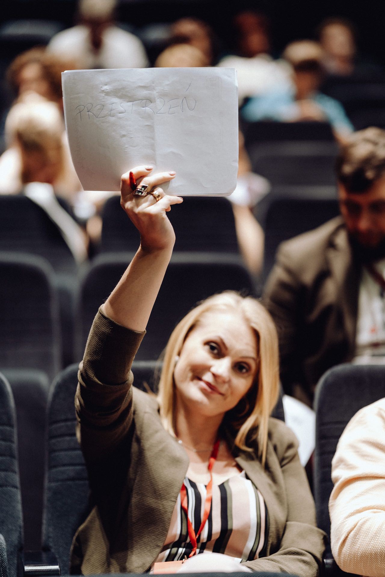 A woman with blond hair, sitting in a dark room, holds a piece of paper with "PRESENT" written on it. She wears a khaki jacket and a lanyard around her neck. Other people are sitting in the background, some of them blurred. Captured by the event photographer warszawa, the image exudes authenticity and commitment.   