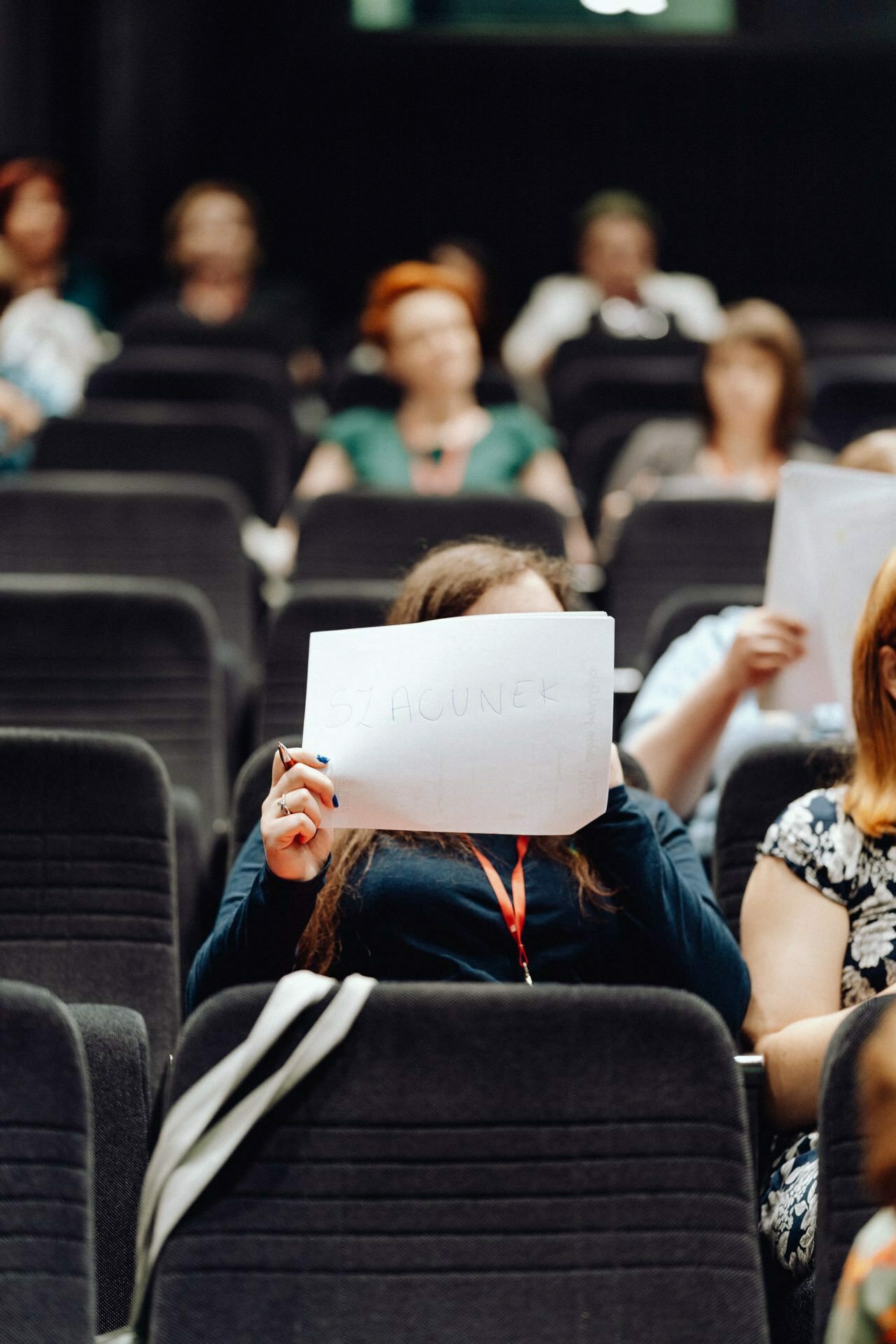 A person in a dark blue top sitting in the audience holds a placard with the handwritten text "HAPPENING" covering his face. Several other people are sitting in the background, some busy talking or looking through documents. The scenery appears to be an event captured by a photographer at an event.  