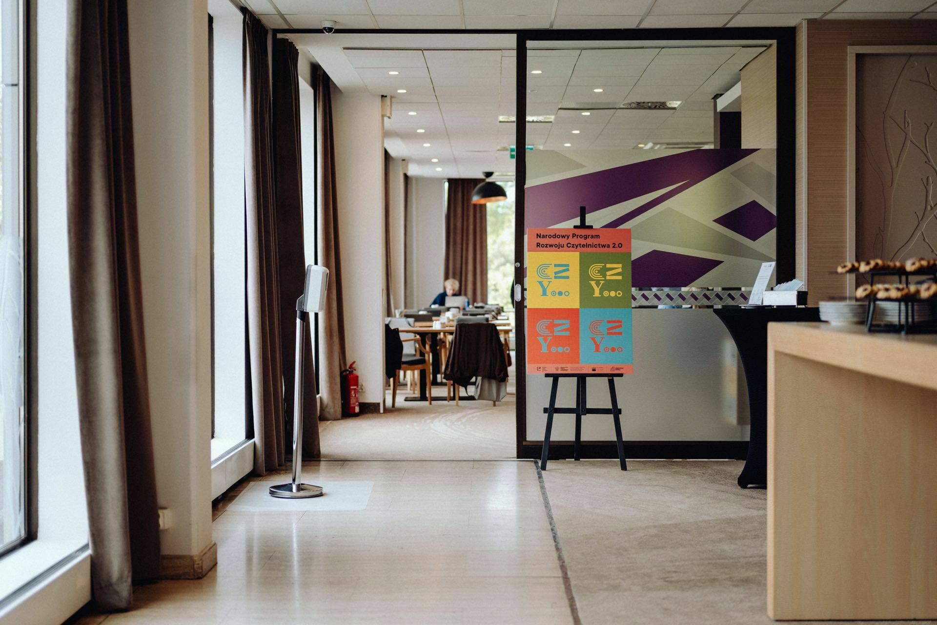 A modern conference room with large windows and curtains. A colorful sign on an easel proclaims: "Presentation Program: E Z Y ABC" with arrows pointing left and right. In the background are tables and chairs, and to the right is a table with drinks, perfect for any session event photographer Warsaw.  