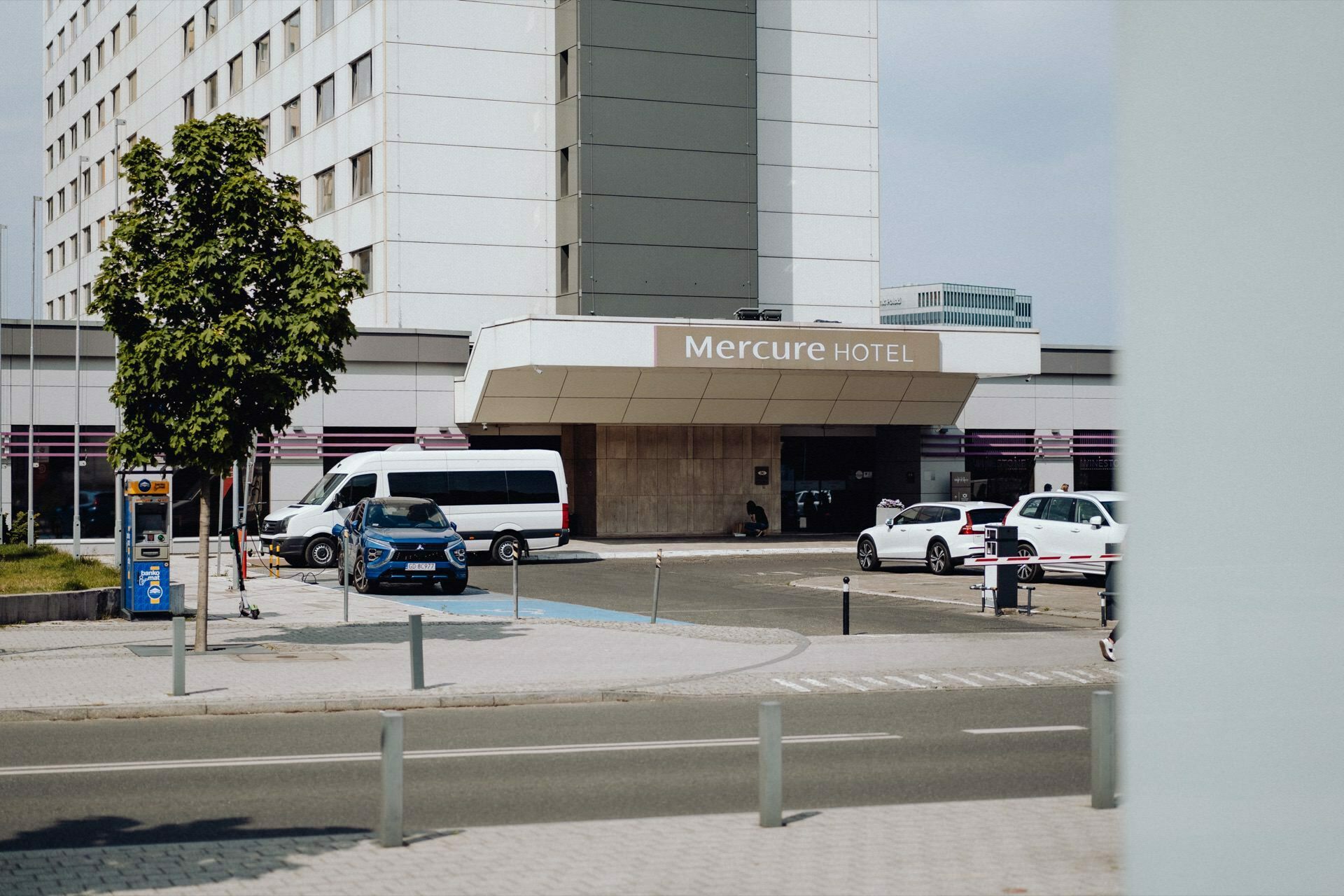 View from the street of the entrance to the Mercure Hotel, which has a modern, multi-story building. A white van and several cars are parked in front of the hotel. The area is surrounded by posts, and there is a tree growing to the left of the entrance, which was clearly captured by a photographer from Warsaw.  