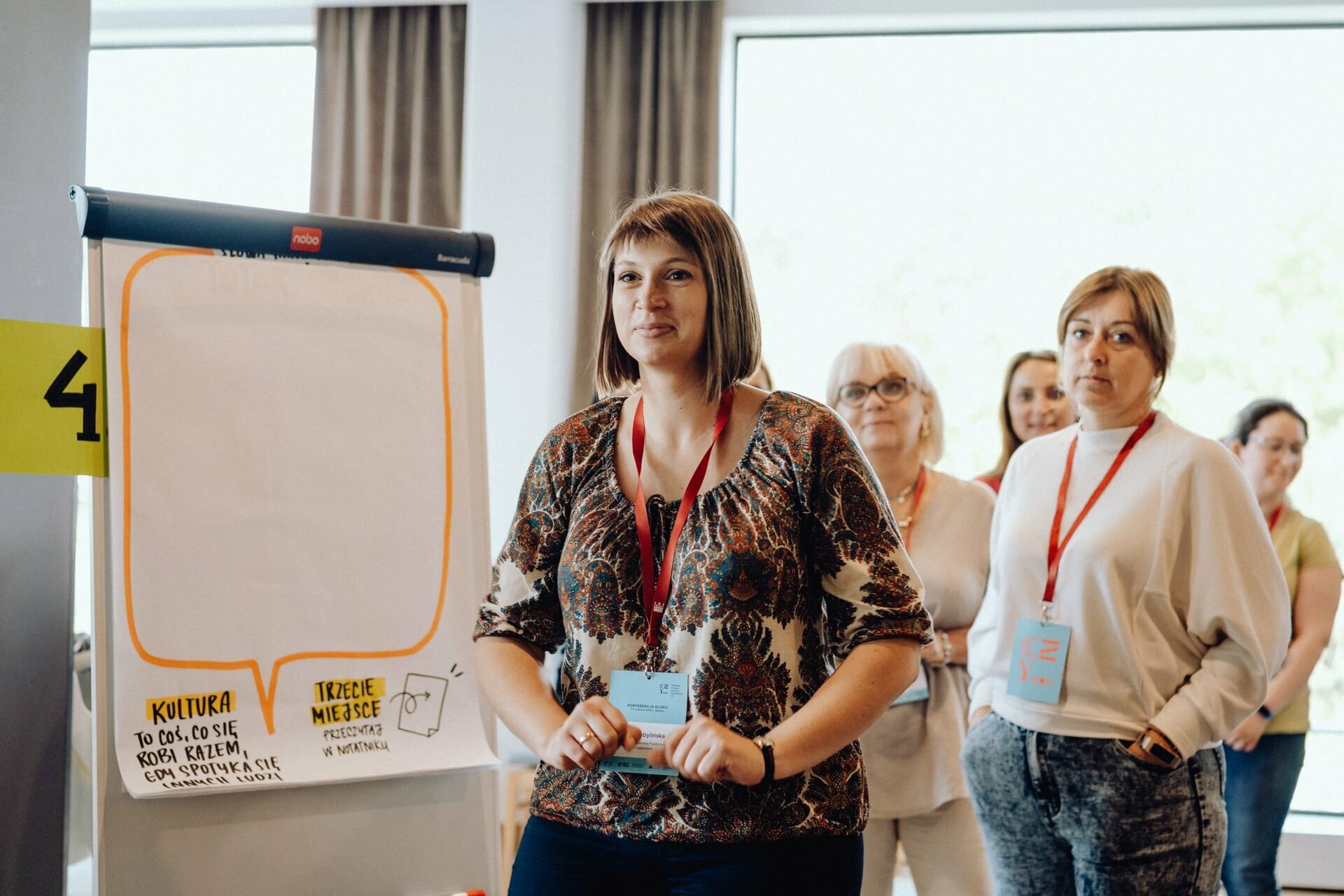 A woman stands in front of the flipchart, followed by three other women, all wearing name badges and lanyards. There are inscriptions and drawings on the flipchart. They appear to be in a conference room with large windows in the background, beautifully captured by a skilled event photographer Warsaw.  