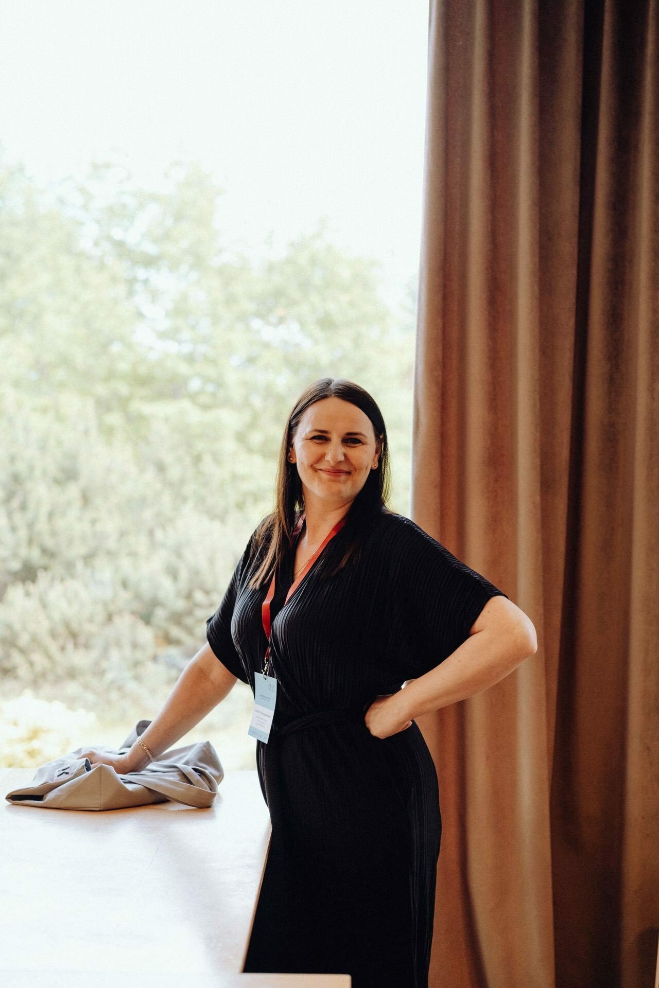 A woman with long, dark hair, dressed in a black outfit and wearing a lanyard with an ID badge, stands in a room by a window, with trees visible outside the window. Smiling slightly, she rests one hand on her hip and the other on a bright surface. Captured by event photographer Warsaw, the moment exudes professionalism.  