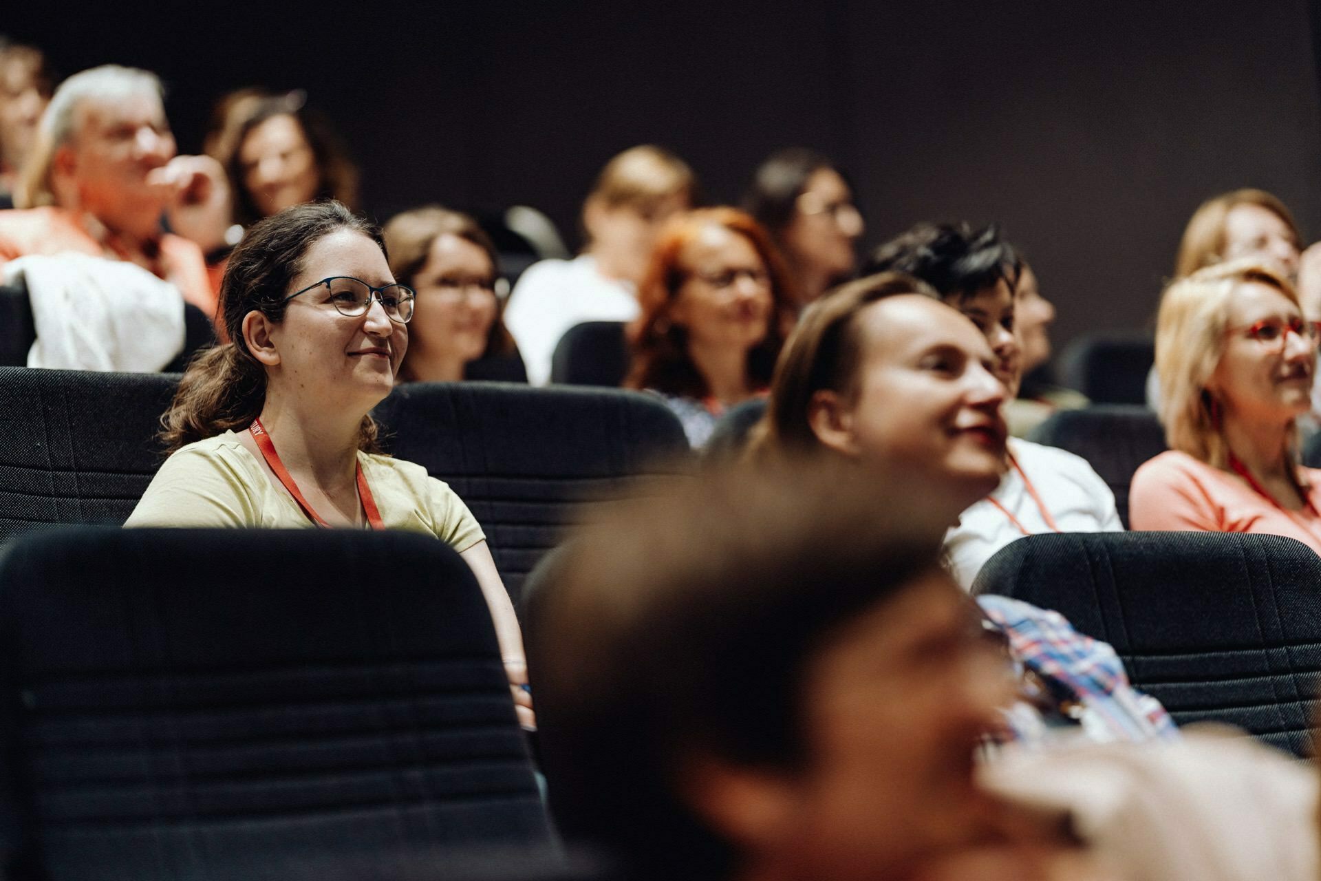 A diverse group of people, mostly women, sit in the rows of a theater or lecture hall. They listen attentively and smile, showing their involvement in the presentation or event taking place. The backdrop is dimly lit, focusing on the audience - an ideal scene for an event photo essay by a photographer from Warsaw.  