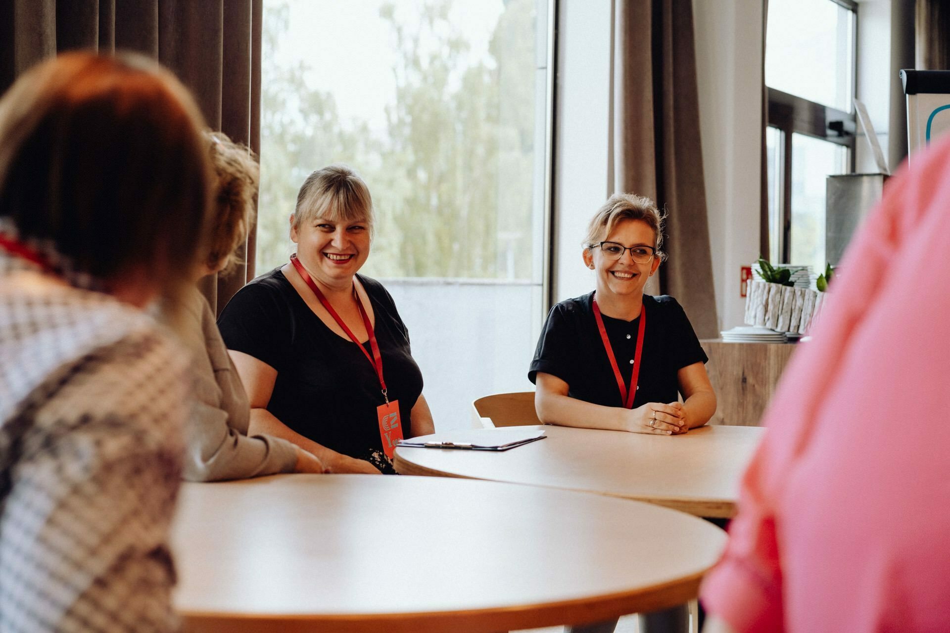 A group of people have gathered around tables in a well-lit room. Two people, one in a black shirt and the other in a dark gray shirt, are smiling and sitting facing the camera with red lanyards around their necks. Other people are partially visible in the foreground, captured by a talented photographer for an event from Warsaw.  