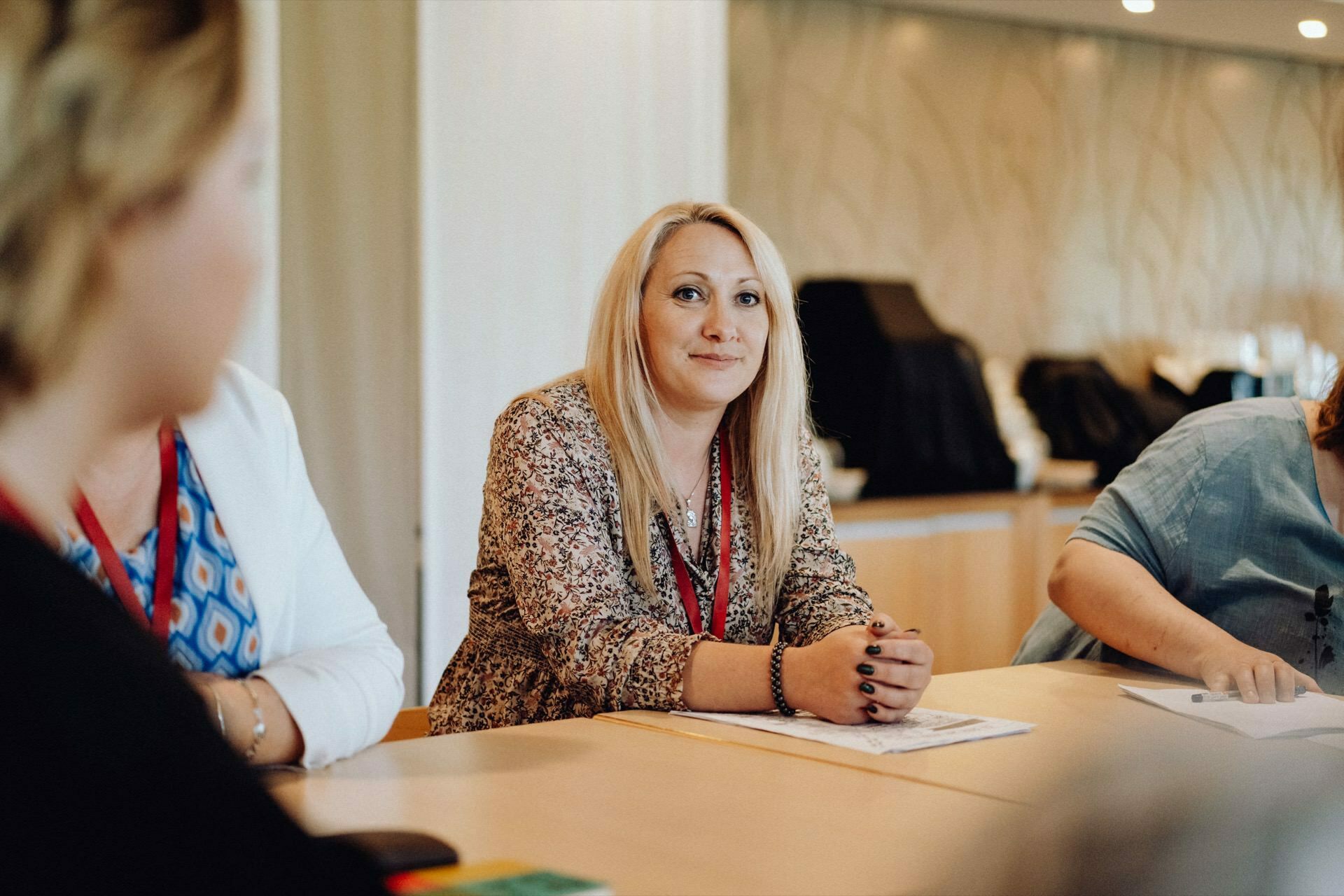 A woman with long blond hair, wearing a floral blouse, sits at a conference table and looks into a camera captured by a photographer at an event. Two other people are partially visible, one in a blue blouse and the other in a white jacket. The background is a modern, well-lit room with beige walls.  