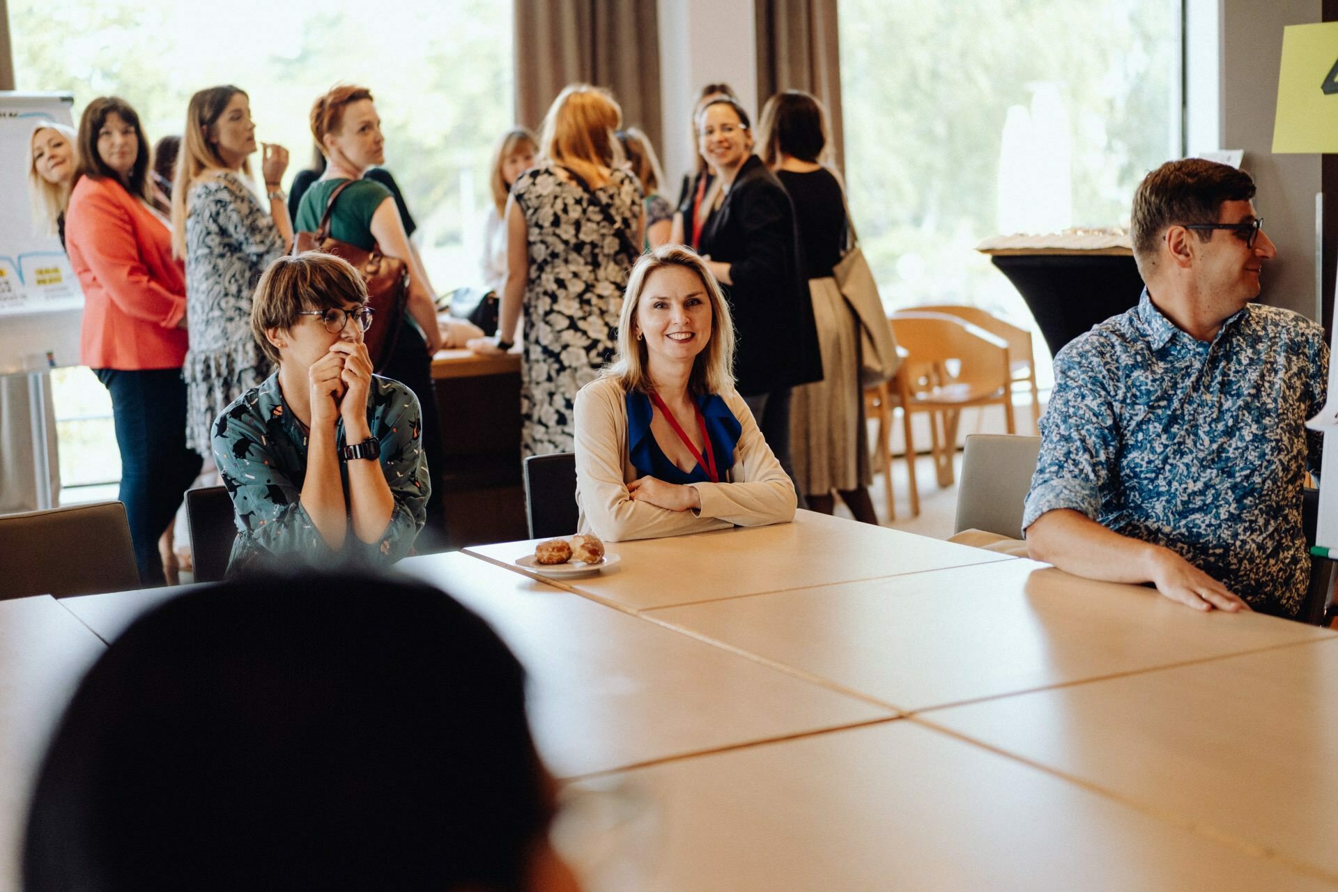 A group of people sit around a table in a brightly lit room, talking and smiling. Some people are eating, others are interacting with each other. Large windows in the background let in natural light, capturing a perfect photo-op of the event.  