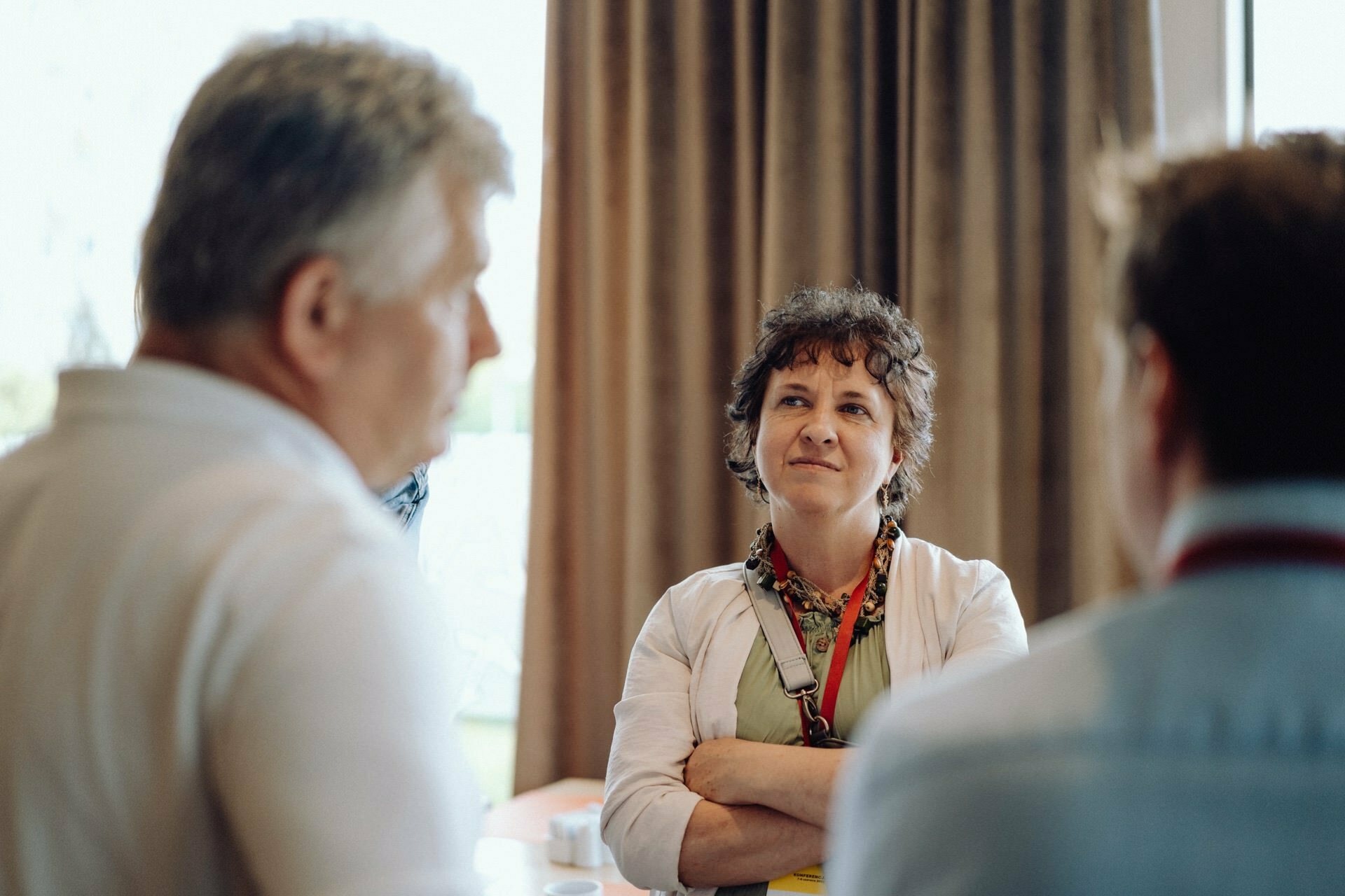 A woman with short curly hair, wearing a light-colored outfit and a colorful headscarf, stands with her arms folded and listens intently to a conversation between two men in white shirts in a room with tall brown curtains in the background - a moment perfectly captured by the photographer at the event.