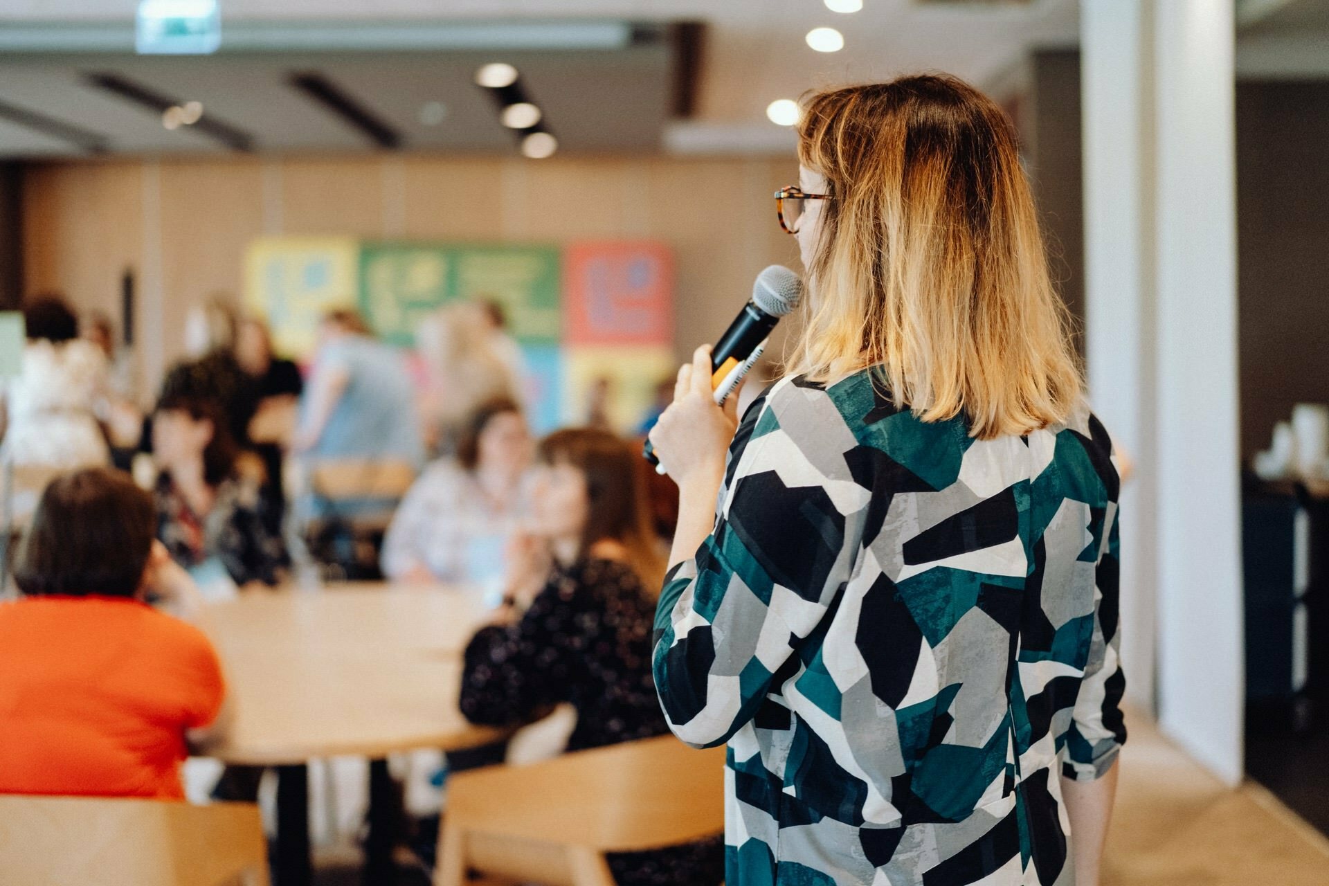 A person holds a microphone and speaks to a group seated in a room with a casual conference-like atmosphere. The audience seems engaged, sitting at tables with a blurry background highlighting the speaker, which was perfectly captured by the photographer for the event. 