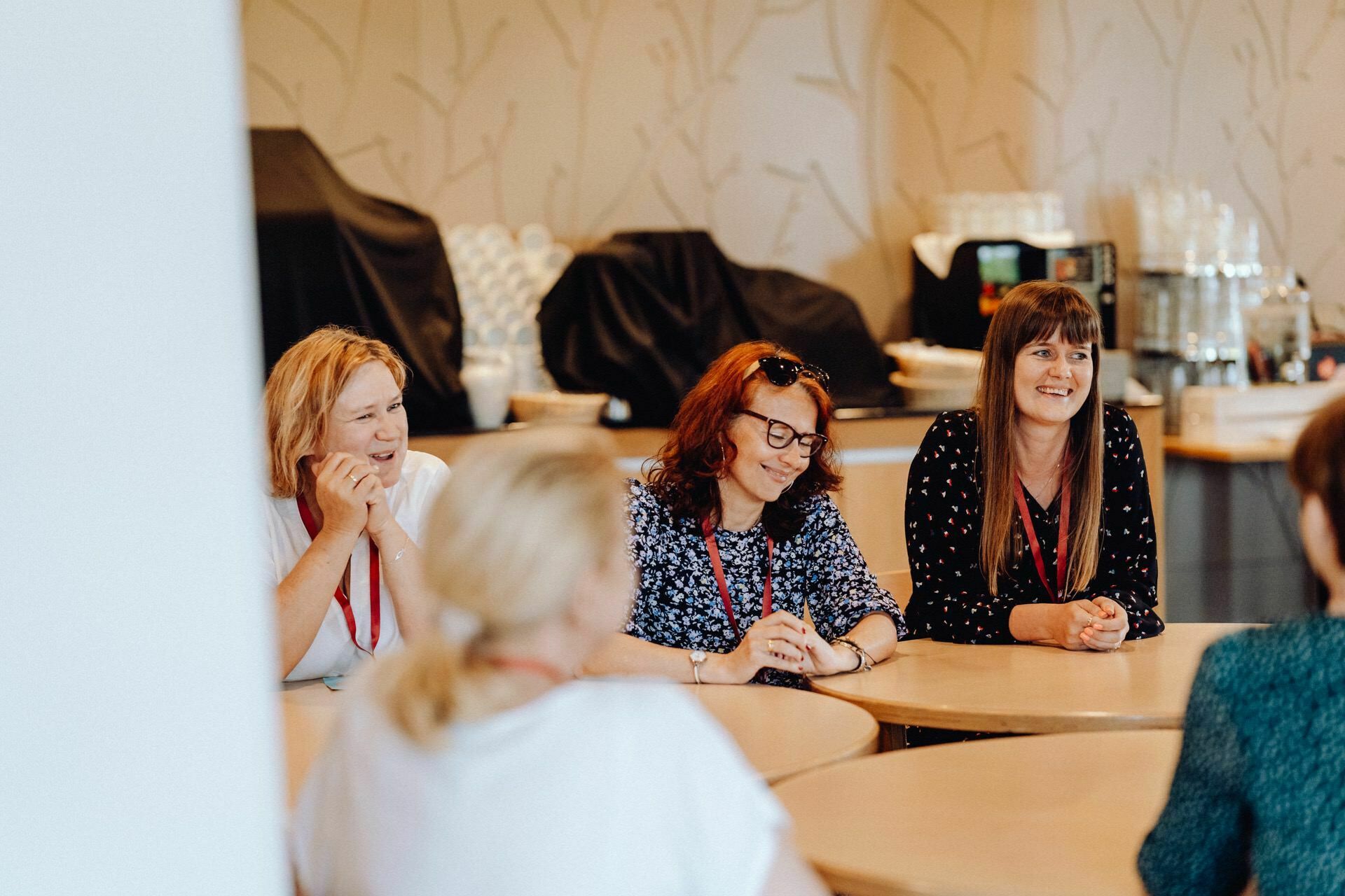 Four women are seated around a table in a casual atmosphere, engrossed in conversation. Three face the camera, one has her back turned. They wear everyday clothes and red lanyards. Shelves and kitchen equipment are visible in the background, beautifully captured by event photographer Warsaw.   