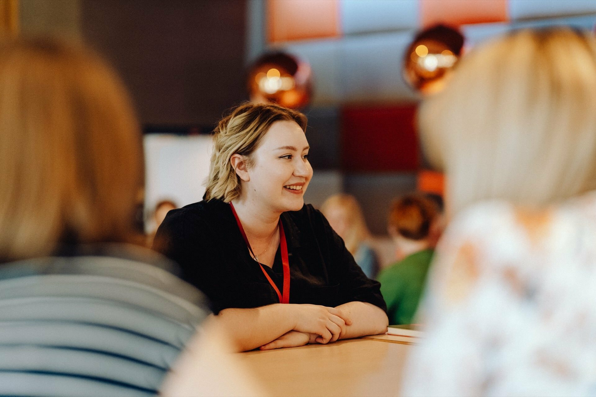 A smiling woman with blonde hair, wearing a black shirt and red lanyard, sits at a table busy in conversation with two other women, whose faces are not visible, beautifully captured by the photographer for the event. The background is blurred, with warm lighting fixtures and modern decor. 