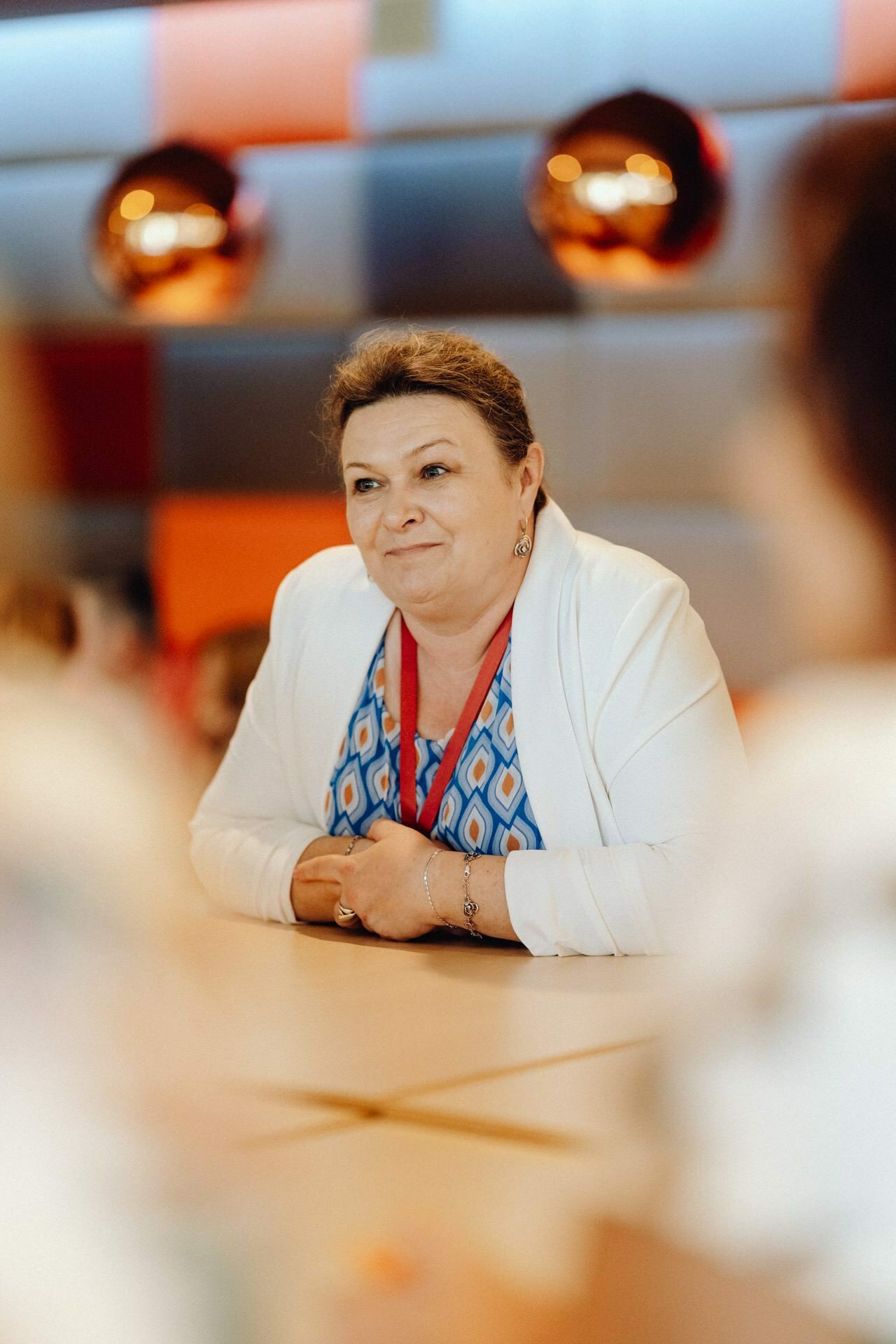 Sitting at a table is a person with short hair, wearing a white jacket imposed over a patterned blue top. The backdrop features colorful, modern decor with metal spherical light fixtures. The person is smiling and gives the impression of being preoccupied with conversation, which was perfectly captured in a photo report of the event by a photographer from Warsaw.  