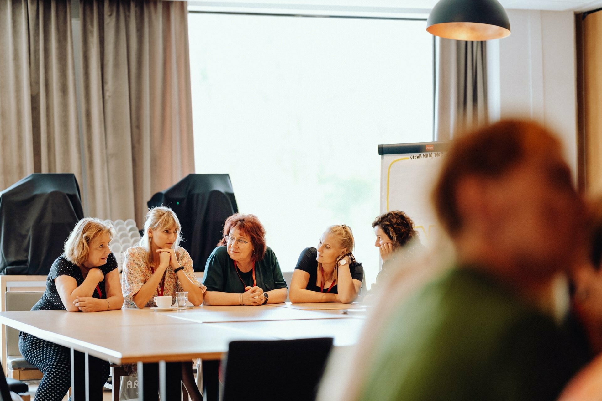 A group of five people sit at a rectangular table in a well-lit room, immersed in conversation. They look thoughtful and focused. The scenery, skillfully captured by the photographer for the event, includes a large window with curtains, a flipchart and a modern pendant lamp. Other people are slightly blurred in the foreground.   