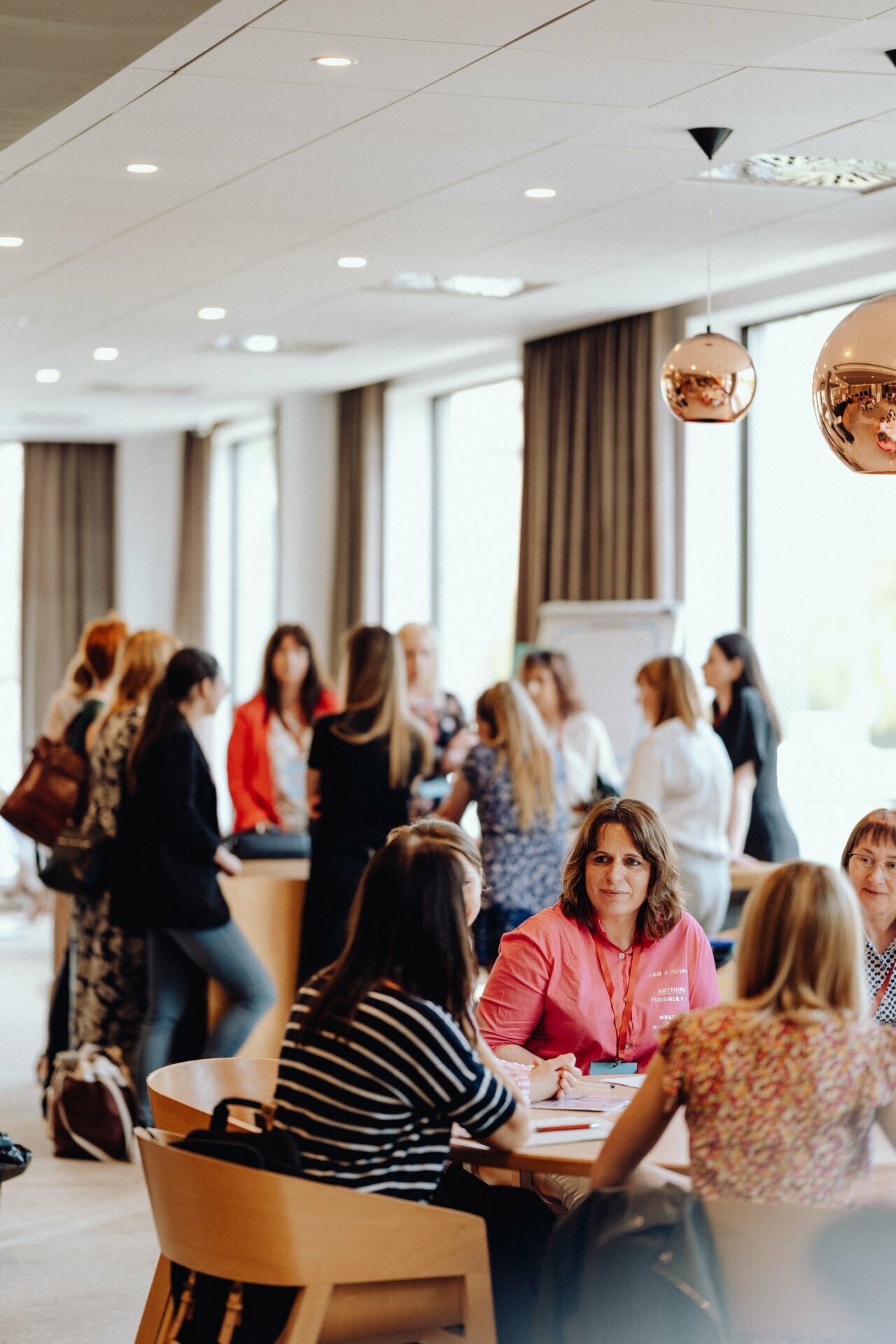 A brightly lit room where people are having conversations. A few women sit at a round table and discuss, while more people stand and talk in the background. The modern decor with large windows and pendant lights creates a scene, perfect for a photographer at an event to capture the event.  