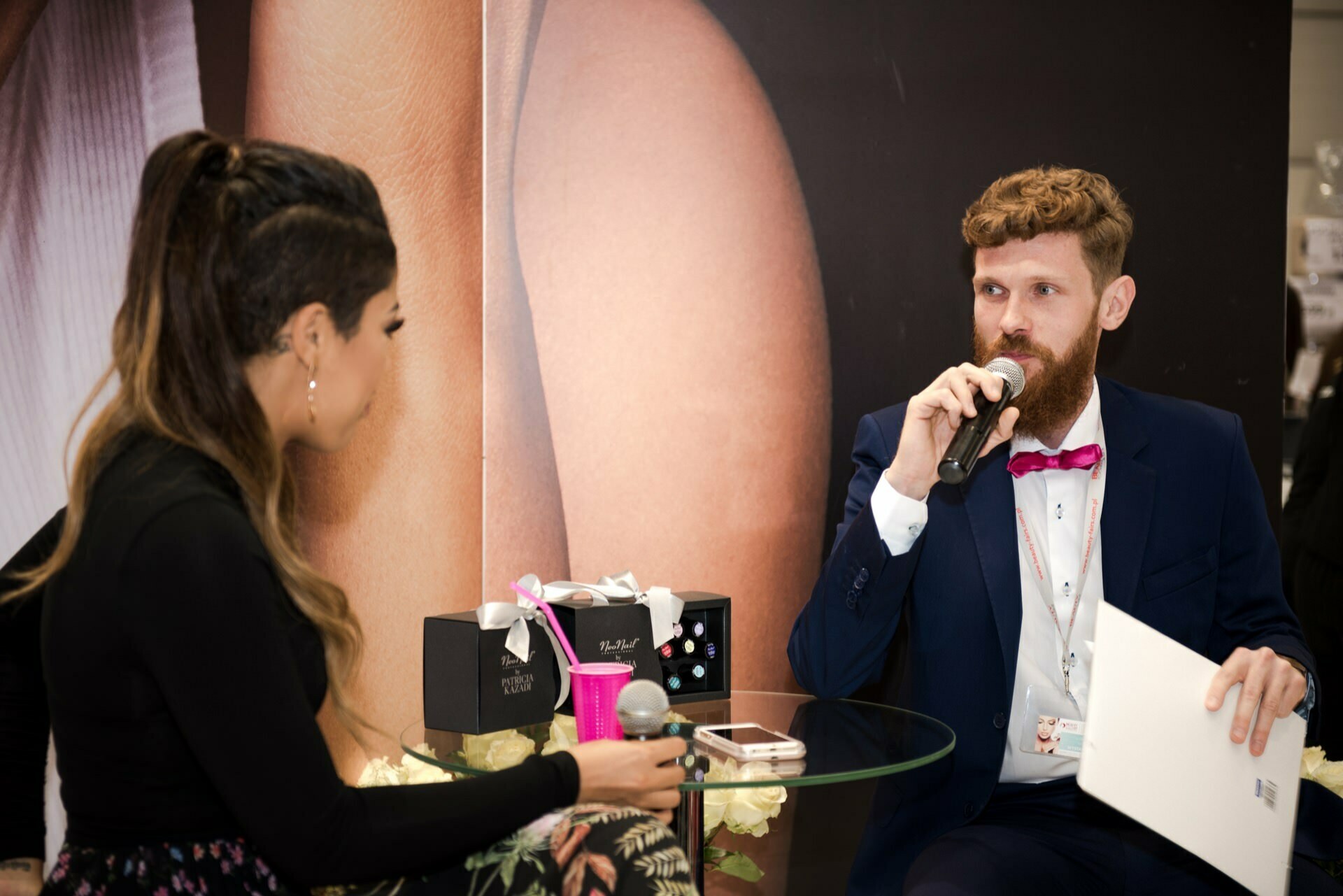 A man with a beard, wearing a blue suit and pink bow tie, speaks into a microphone while holding a newspaper. He is sitting at a round glass table with gift boxes, presumably for the Beauty Forum. Opposite him sits a partially visible woman, holding a microphone. Behind them is a large photographic background.   