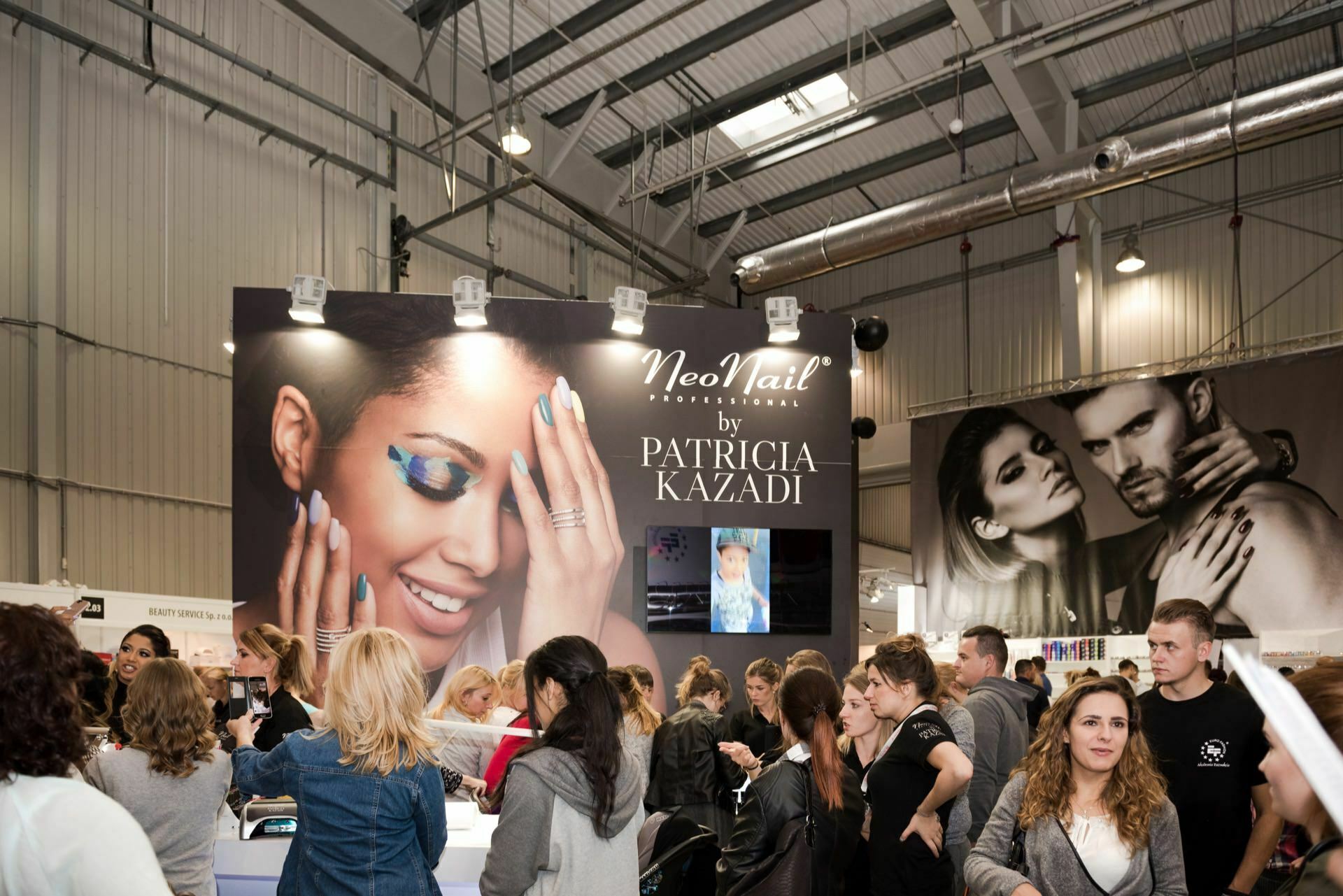 A bustling cosmetics fair where people gathered around a booth with large NeoNail signs by Patricia Kazadi. The ad shows a smiling woman with expressive, artistic eye makeup. Captured by event photographer Marcin Krokowski, the venue is an industrial-style hall with high ceilings and exposed ceiling beams.  