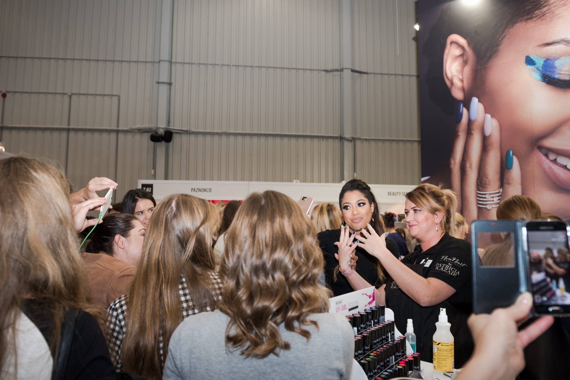 At the event, a large group of people, mostly women, gather around a cosmetics stand. Two women in the middle interact with the crowd, one holding a bottle of nail polish. Behind them is a poster showing a close-up of a woman with colorful eye makeup. Captured by Marcin Krokowski, event photographer.   