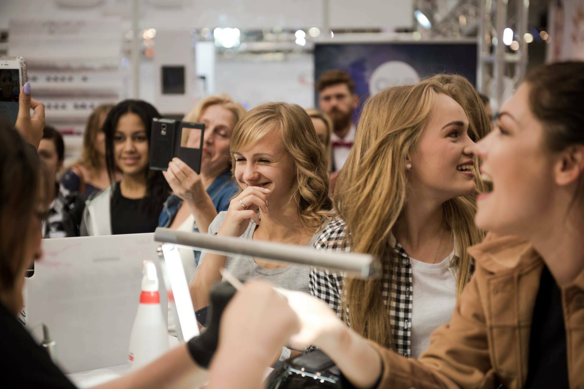 A group of people, mostly young women, gather in a brightly lit room. They are smiling, laughing, and some are taking pictures with their phones. The lively and cheerful mood is reminiscent of an event or meeting captured by prominent Warsaw photographer Marcin Krokowski.  