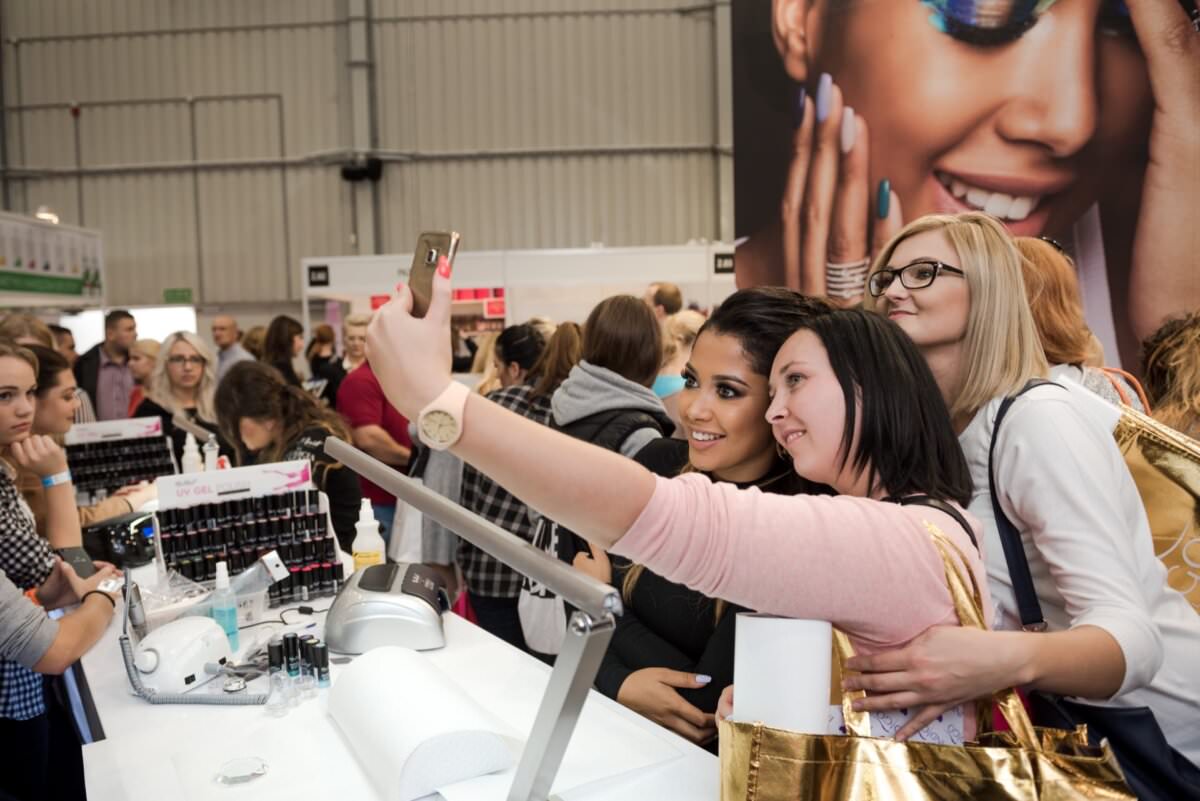 Grupa trzech kobiet robi sobie wspólne selfie na ruchliwej wystawie lub konwencji. Stoją przy stoisku z różnymi produktami, w otoczeniu innych uczestników. W tle widoczna duża reklama z uśmiechniętą kobietą w makijażu, doskonale uchwycona przez Marcina Krokowskiego, zdolnego fotografa eventowego.