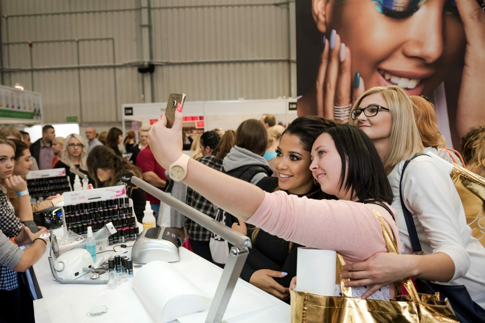 A group of three women take a selfie together at a busy exhibition or convention. They are standing at a booth with various products, surrounded by other attendees. A large advertisement with a smiling woman in makeup is visible in the background, perfectly captured by Marcin Krokowski, a skilled event photographer.  