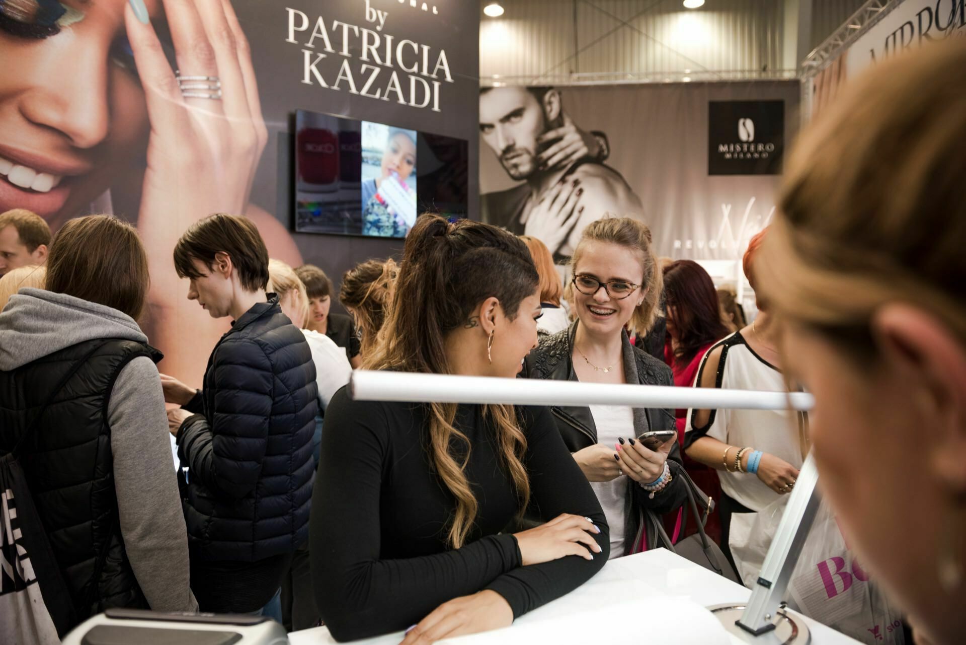 A group of people gather at a booth in the exhibition hall. Most are facing a woman sitting at a table with salon equipment. Behind her is a large advertisement showing two models and the text "author: Patricia Kazadi." Photographer Marcin Krokowski captures the event as some people use their phones.   