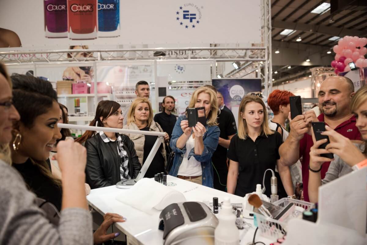 Grupa ludzi gromadzi się wokół stoiska na konwencji lub targach. Robią zdjęcia i filmy telefonem, zupełnie jak na imprezie ze znanym fotografem eventowym Marcinem Krokowskim. Na stoisku w tle eksponowane są produkty oraz banery. Atmosfera jest żywa, a uczestnicy zaangażowani i uśmiechnięci.