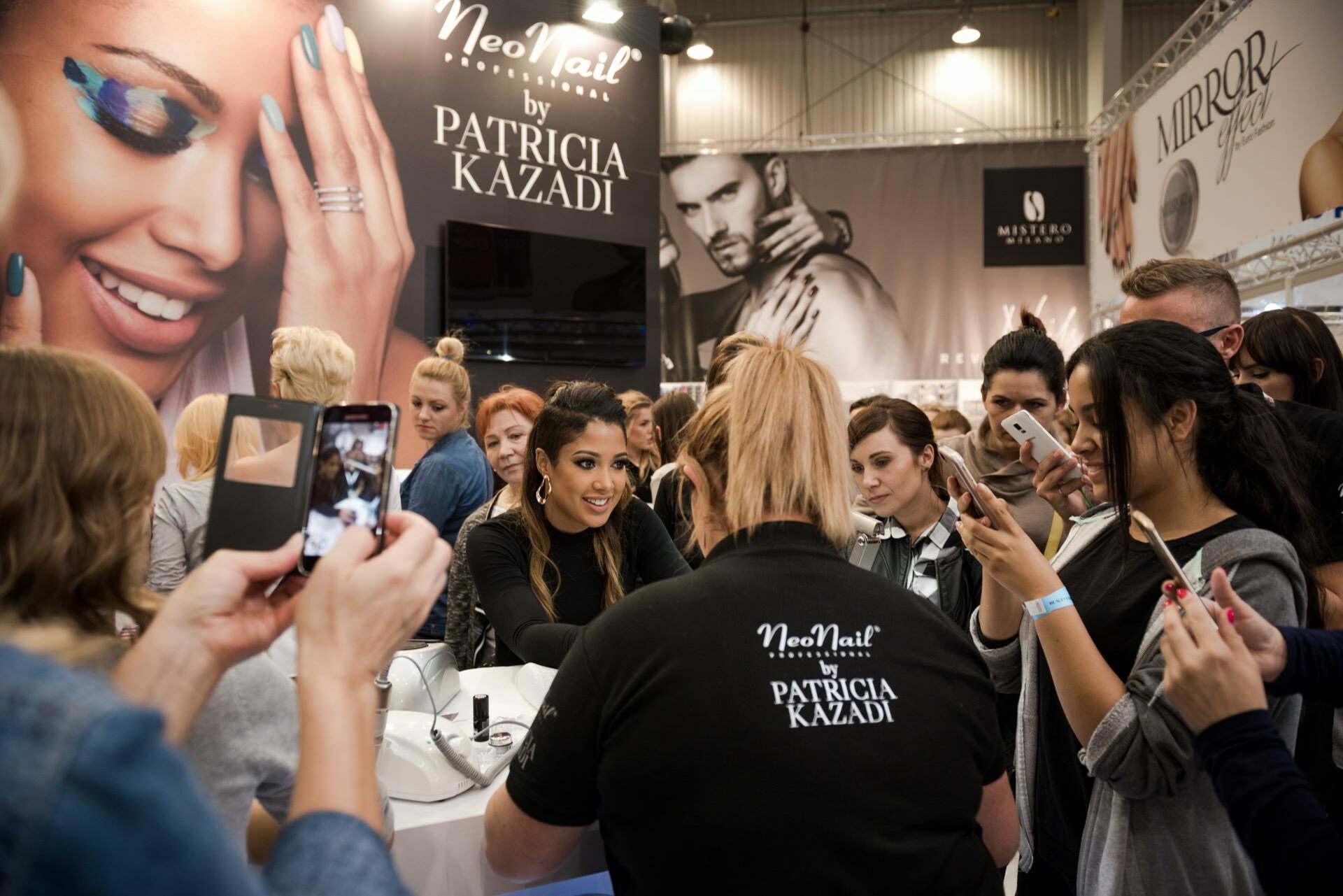 A large group of people gather at a booth with "Patricia Kazadi" and "NeoNail" banners. The main woman, presumably Patricia Kazadi, smiles as she talks to fans and hands out autographs. Marcin Krokowski, an event photographer from Warsaw, captures the bustling scene as several people take photos or videos with smartphones.  