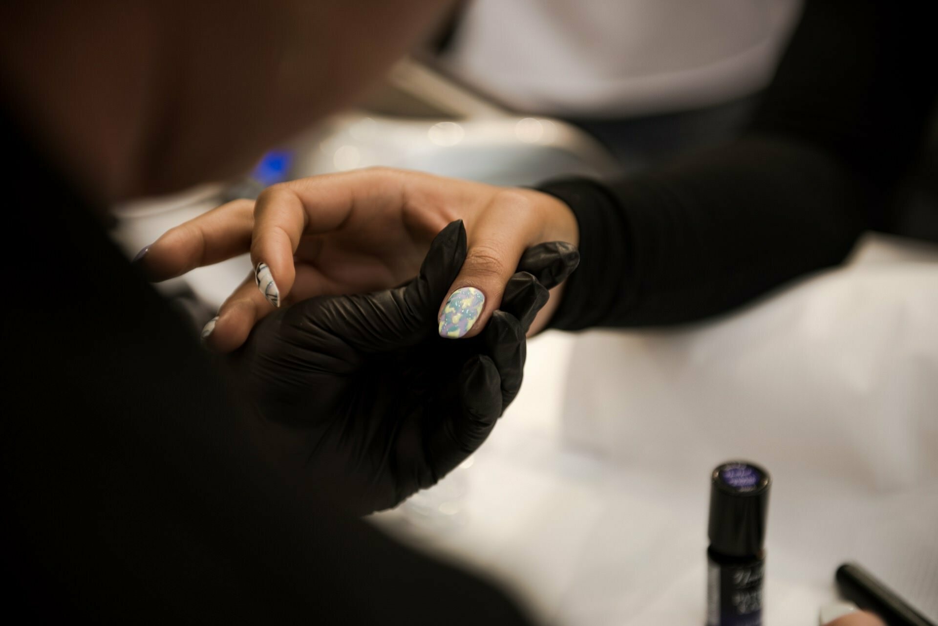 A close-up of a person undergoing a manicure treatment by Marcin Krokowski, a well-known photographer in Warsaw. The manicure person puts on black gloves and gently holds the client's hand, exposing the iridescent nail pattern. Various manicure tools and products are visible in the background.  