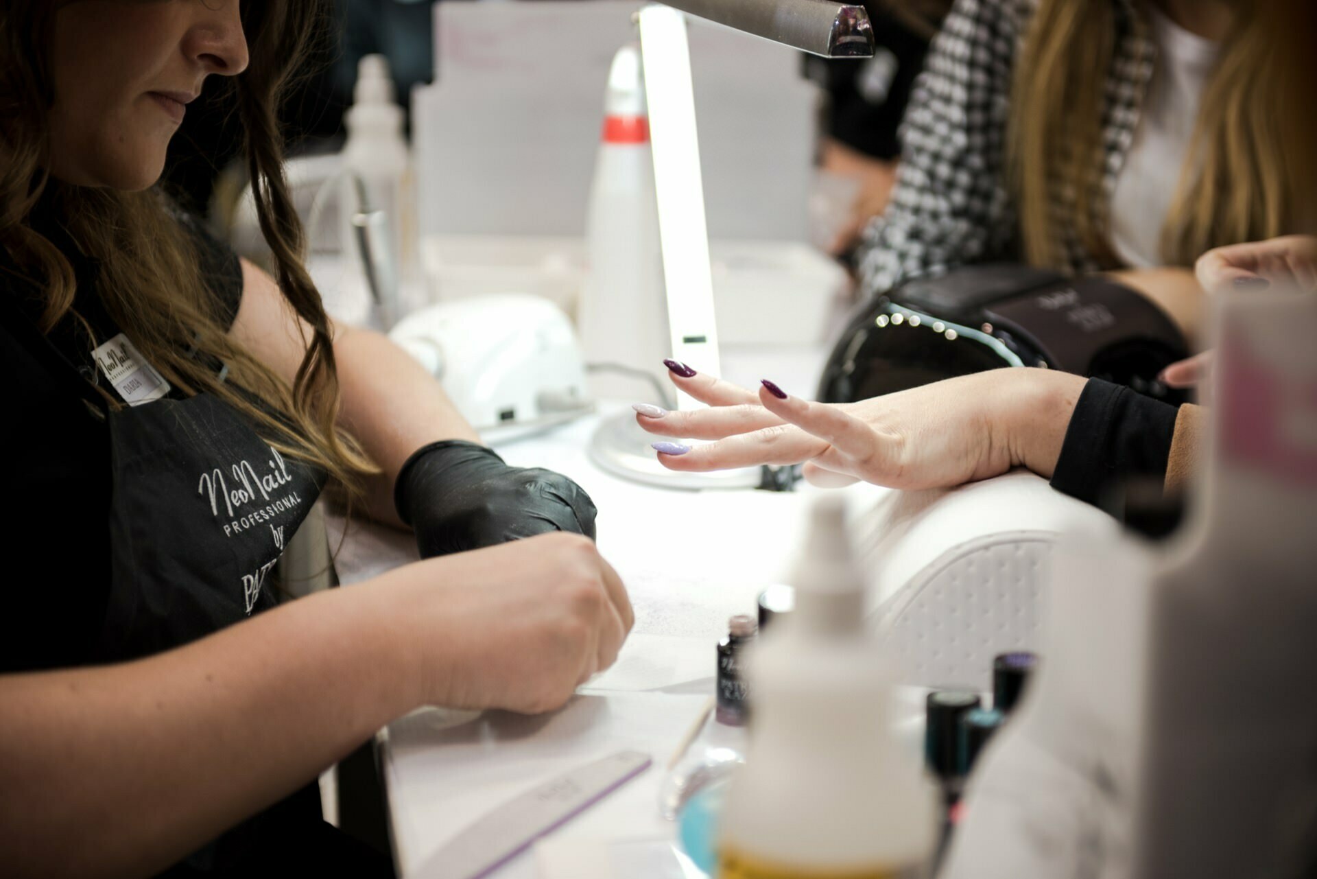 A woman sits at a table while a nail stylist dressed in a black apron and black gloves does her nails. Various bottles and nail utensils lie on the table, beautifully captured by well-known photographer in Warsaw, Marcin Krokowski. Another person in the background is also getting a manicure.  