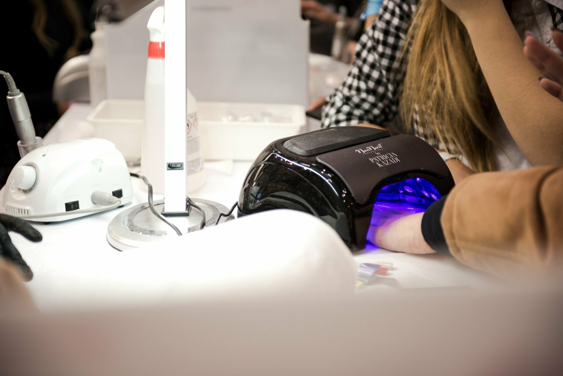 A person giving a hand manicure under a UV nail lamp on a white table. Various manicure tools are visible, including an electric nail cutter and a desk lamp. Another person's arms are partially in the frame helping with the manicure - beautifully captured by photographer in Warsaw, Marcin Krokowski.  