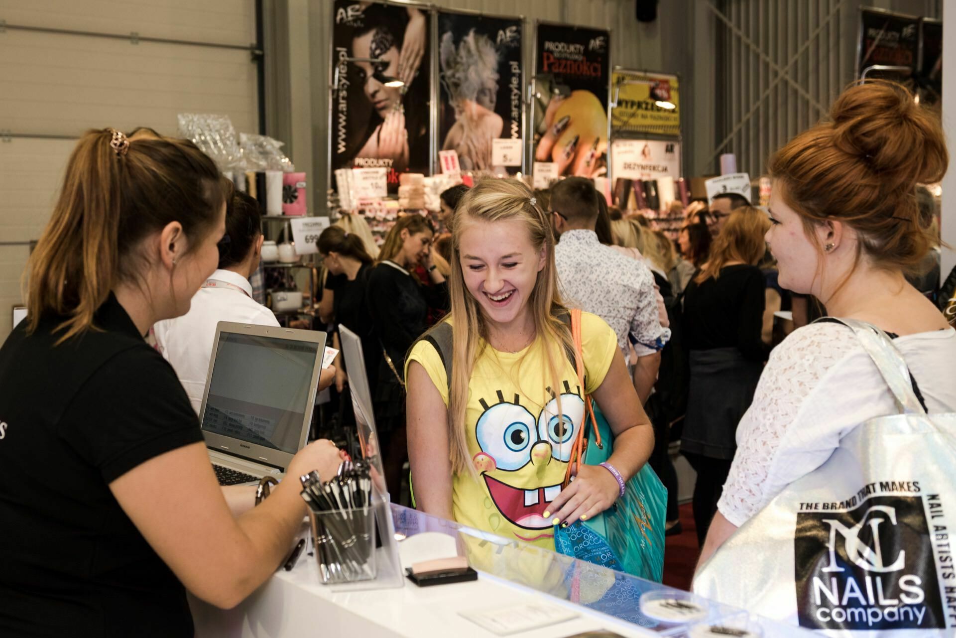 A woman in a SpongeBob SquarePants T-shirt smiles as she talks to a representative at the booth. The booth captured by Marcin Krokowski is bustling with various displays of cosmetics. Nearby, another visitor with a branded bag watches the developments.  