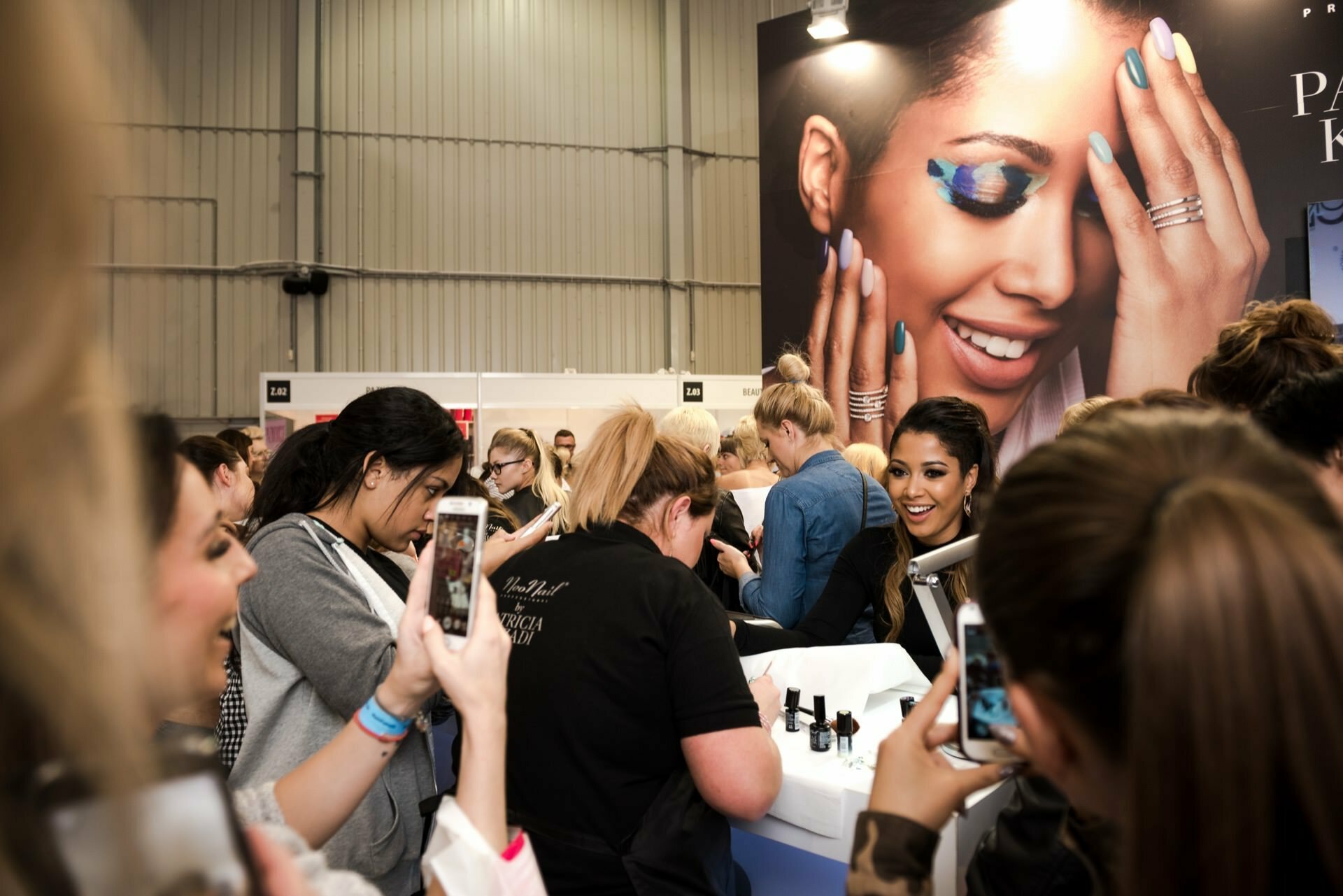 A crowd gathers around a makeup demonstration booth at a beauty event. Many attendees pull out their phones and take photos and videos. A large poster showing a woman with colorful eye makeup is visible in the background, and the whole thing was captured by Marcin Krokowski, a well-known photographer in Warsaw.  