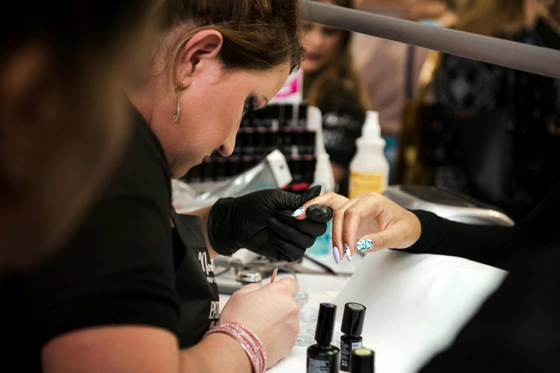 A nail technician wearing black gloves meticulously applies nail polish to a client's nails at a beauty salon. The nails are partially painted white with a blue and white floral pattern. Various nail polish bottles and tools are visible on the table, captured by Marcin Krokowski, a talented photographer in Warsaw.  